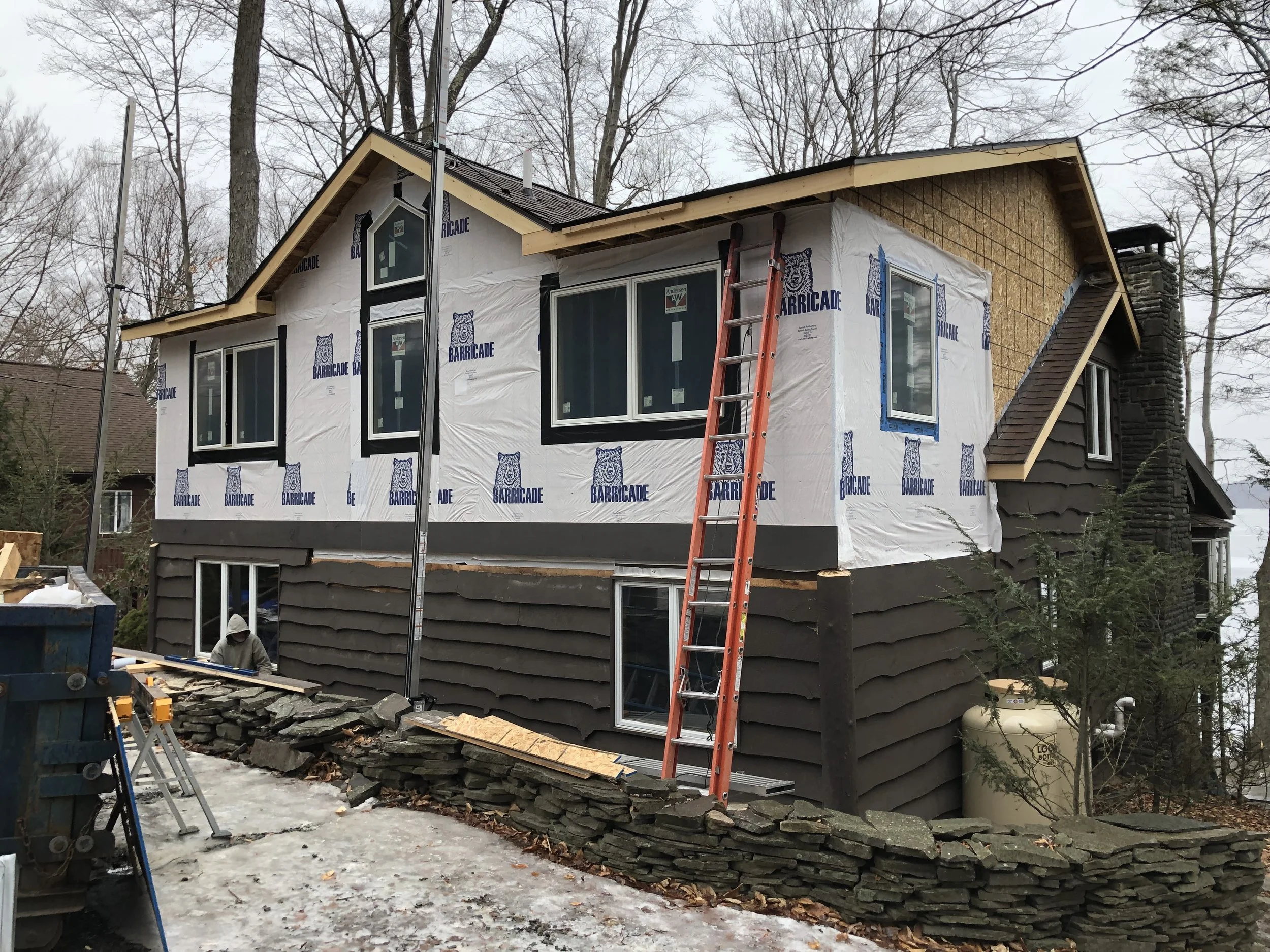 House under construction with insulation and windows installed, orange ladder leaning against the building, workers working nearby, trees in the background, and construction materials on the ground.