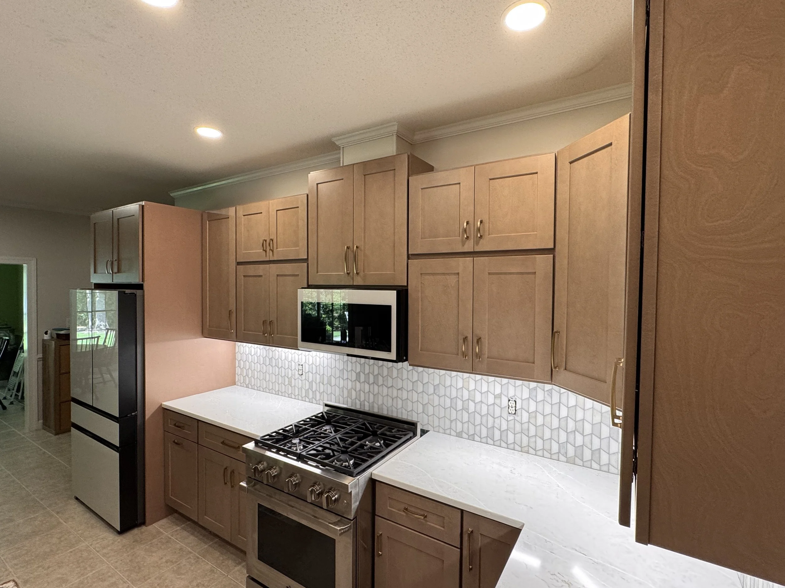 Modern kitchen with brown wooden cabinets, white marble countertops, and a stainless steel stove with a gas cooktop. There is a black microwave built into the cabinetry, a large refrigerator to the left, and a decorative tile backsplash behind the stove.