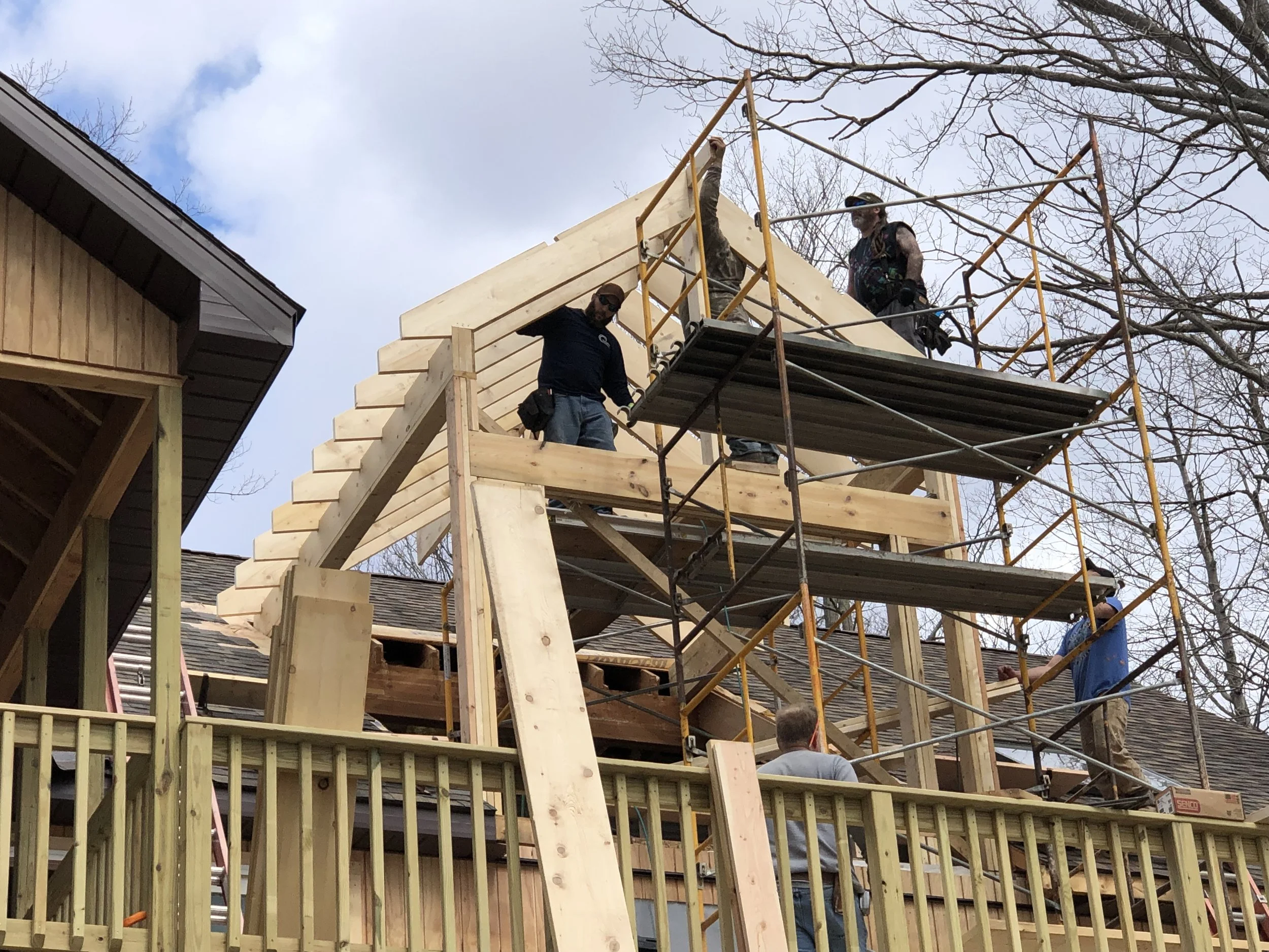 Construction workers building a wooden structure on the roof of a house, using scaffolding and ladders, with trees and partly cloudy sky in the background.