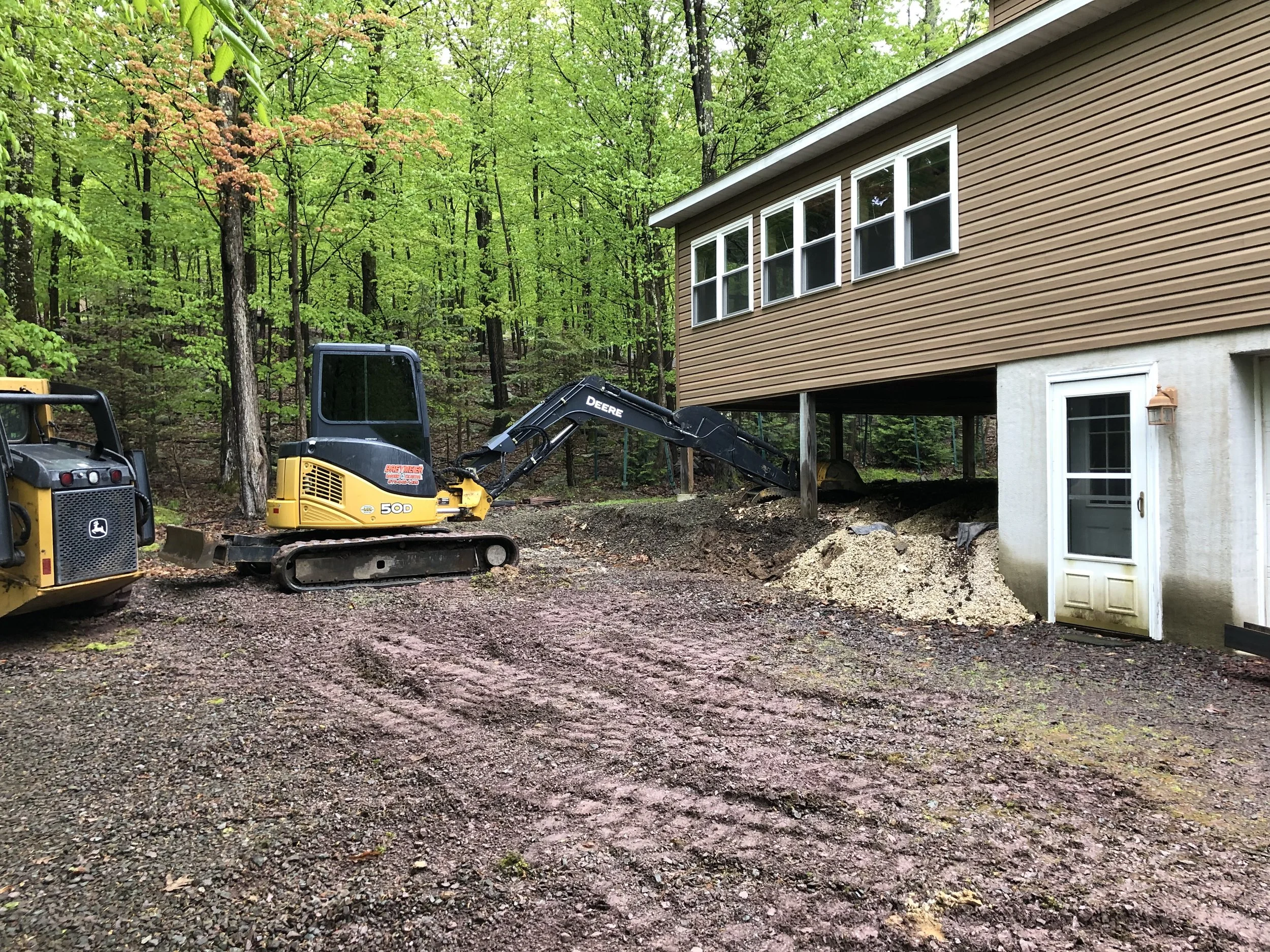 Construction equipment, including a John Deere mini excavator, working on the foundation of a house in a wooded area, with soil and gravel in the foreground.