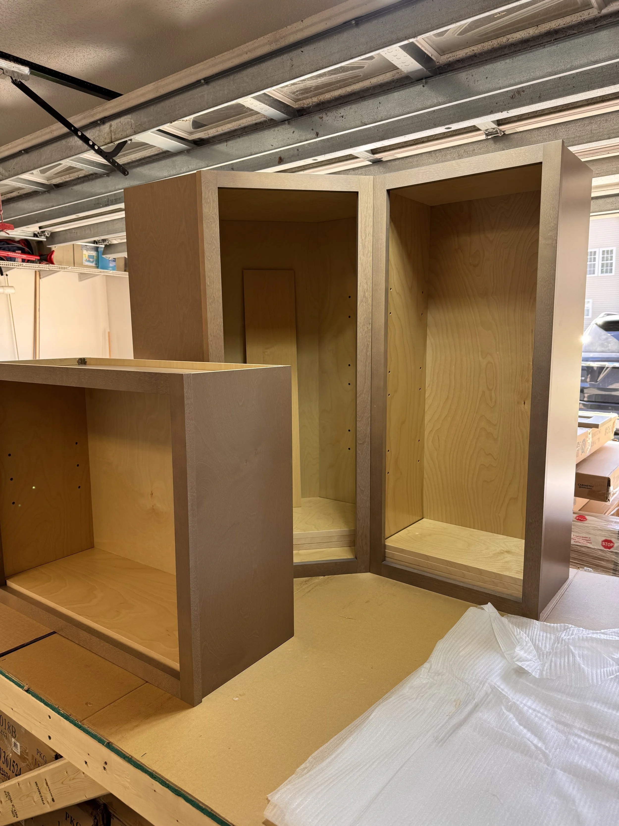 Three unfinished wooden cabinet frames with drilled holes, placed on a worktable in a garage, with packaging and tools in the background.