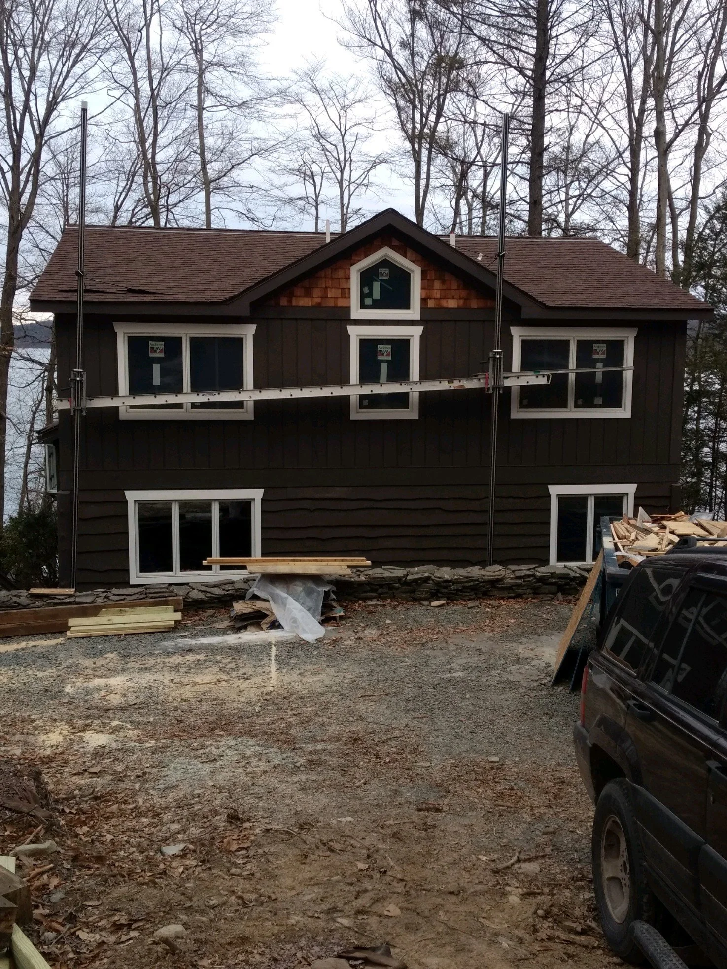 A two-story house under construction with dark brown siding and white window frames, surrounded by leafless trees. Construction materials and a parked vehicle are visible in the foreground.