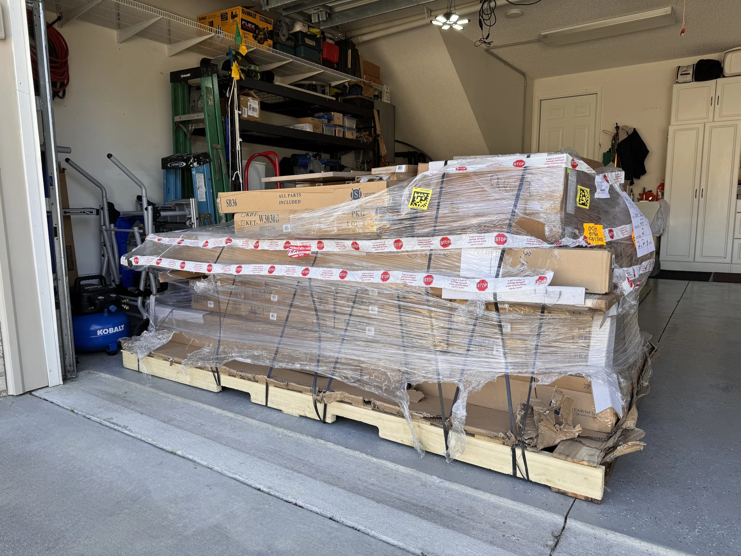 Pallets of boxed and wrapped packages stored in a garage, with tools and storage shelves in the background.
