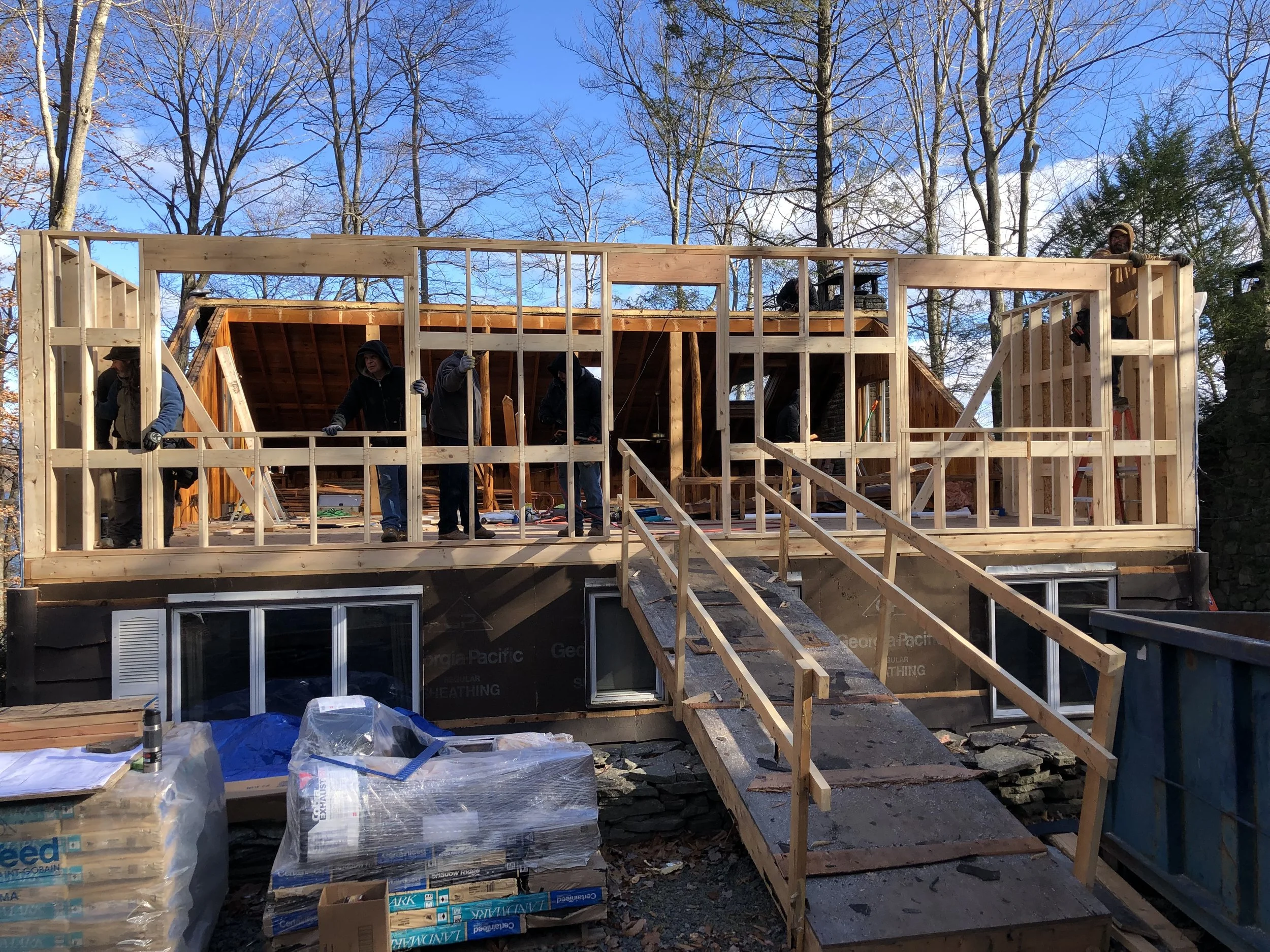 People working on the framing of a house with wooden beams, ladders, and construction materials outdoors with trees and a blue sky in the background.