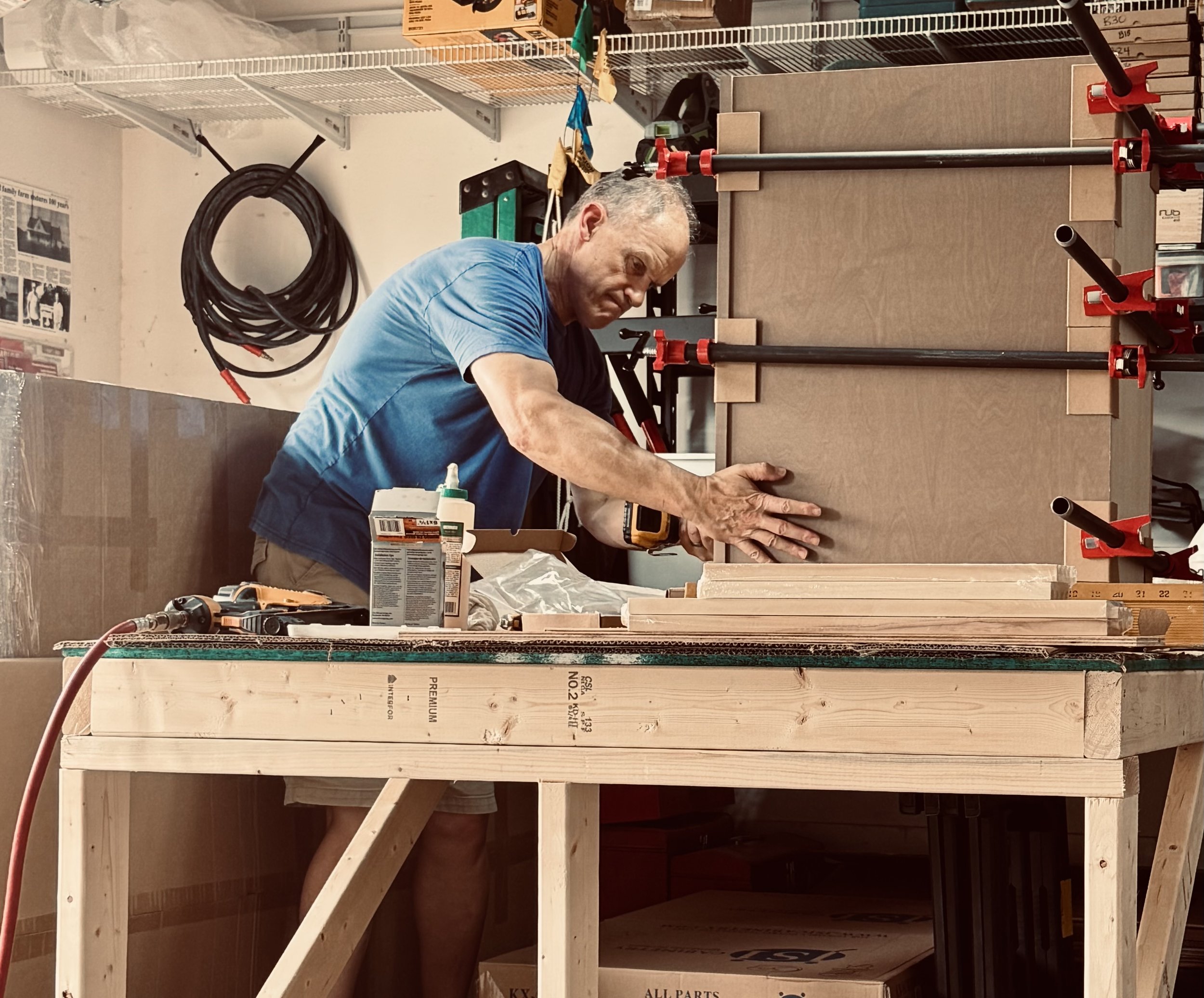 A man working on a woodworking project in a garage, applying glue to a piece of wood with various tools, clamps, and woodworking materials on the table.