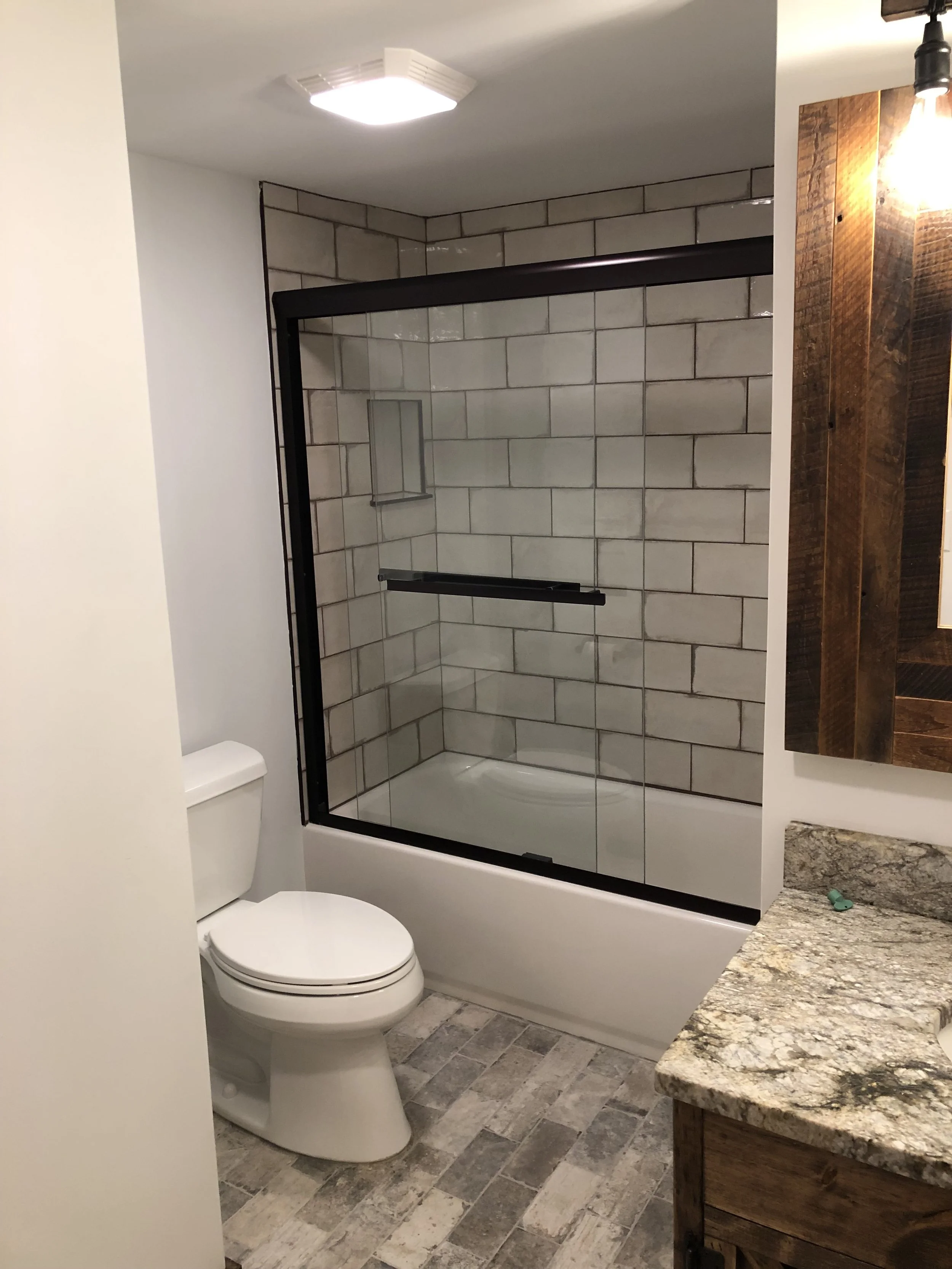 Bathroom with a white toilet, a glass-enclosed shower with beige brick tiles, a granite countertop, and a wooden cabinet. Recessed lighting on the ceiling.