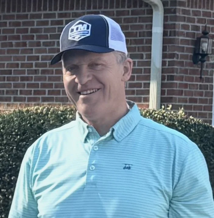 A middle-aged man smiling outdoors, wearing a light blue polo shirt and a navy and white caps with a logo.