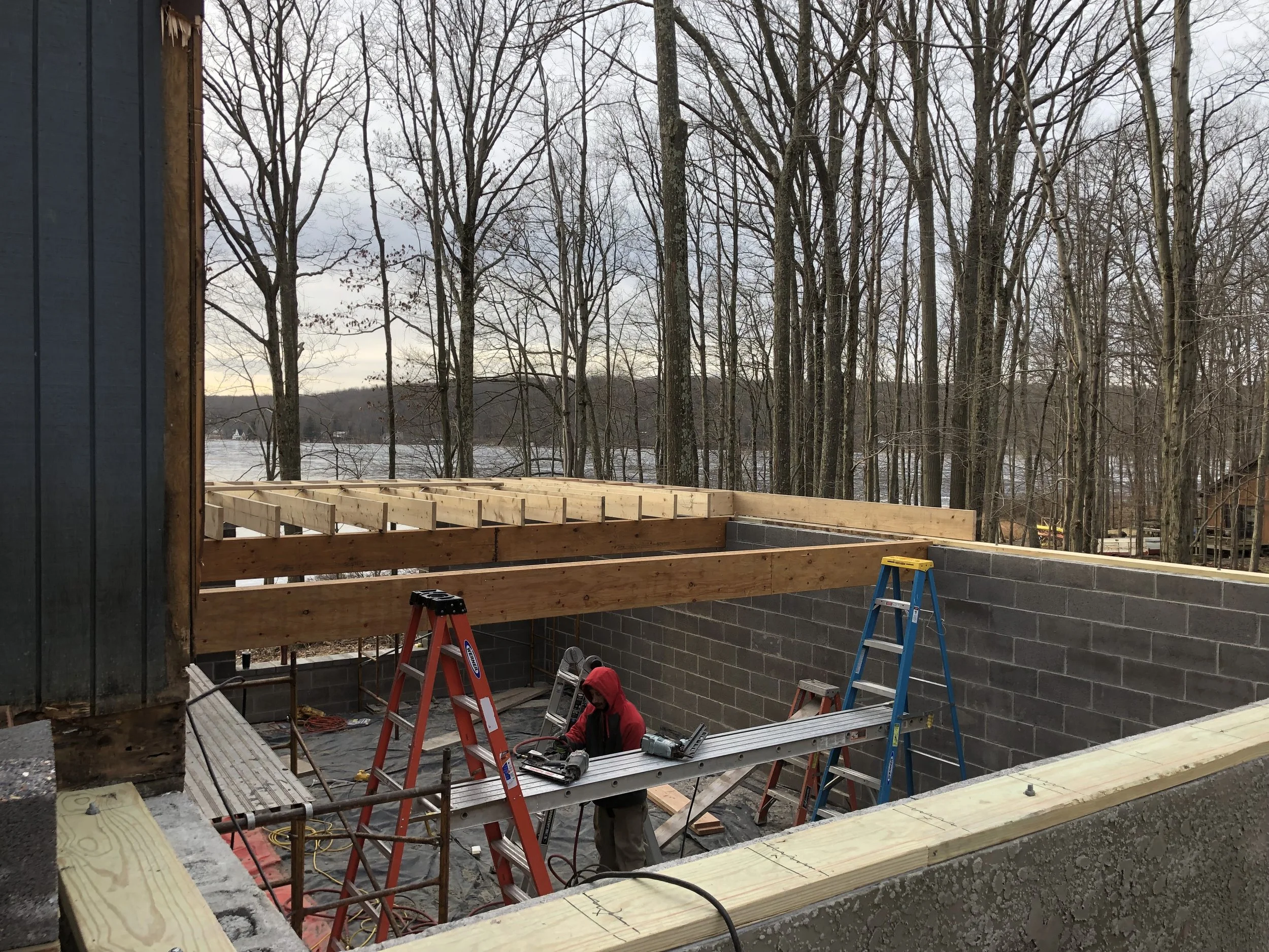 Construction worker working inside a partially built wooden structure during daytime, with trees and a body of water in the background.
