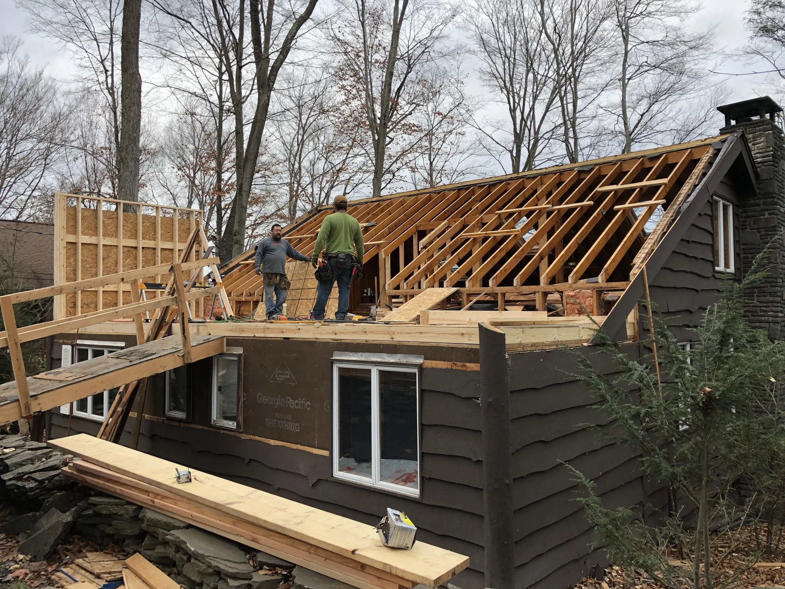 Two construction workers building a new roof on a house, with the roof trusses and framing in progress, surrounded by trees during overcast weather.