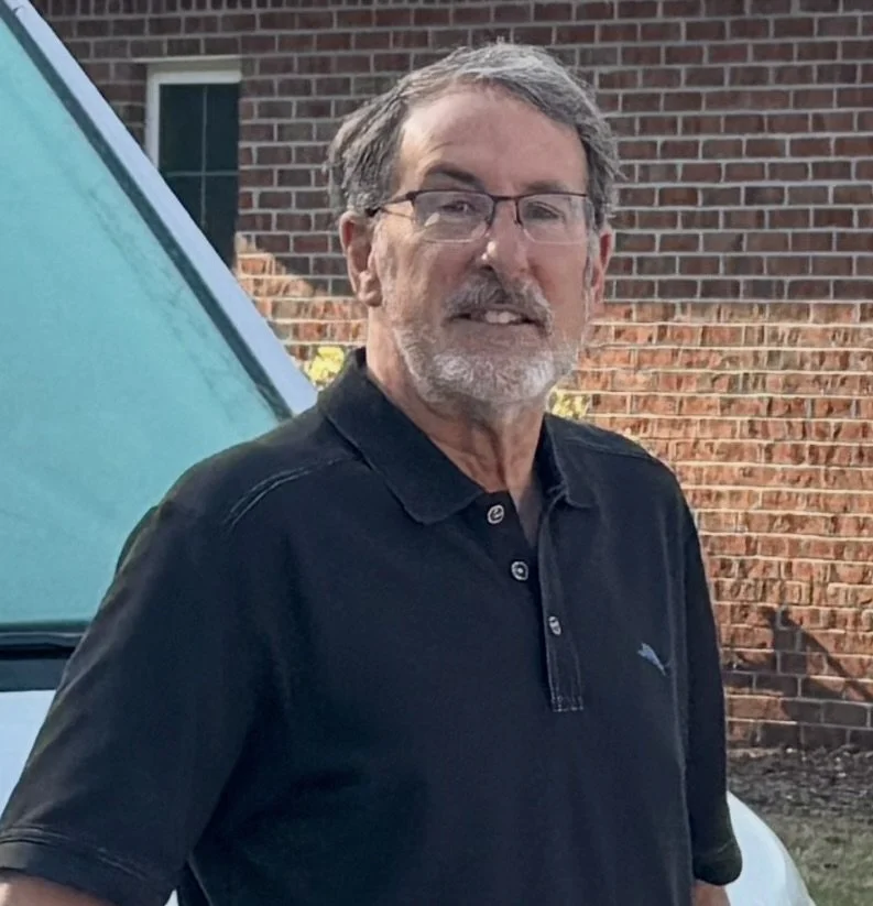 An older man with glasses and gray hair, wearing a black polo shirt, standing outdoors in front of a brick building and a vehicle with a teal-colored hood.