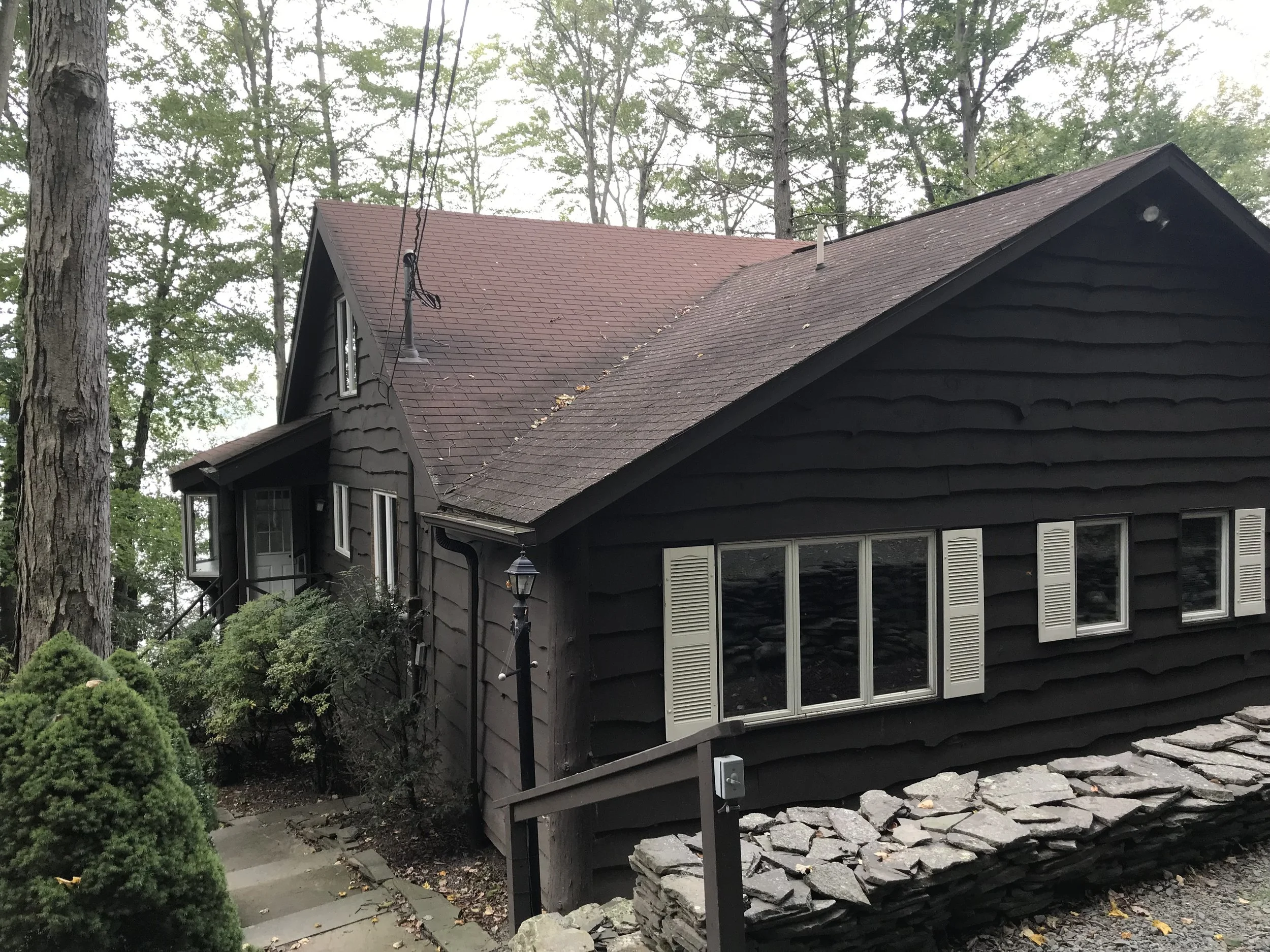 A black wooden house surrounded by trees, with a stone foundation, white shutters on windows, a small front porch with stairs, and a black lamp post near the walkway.