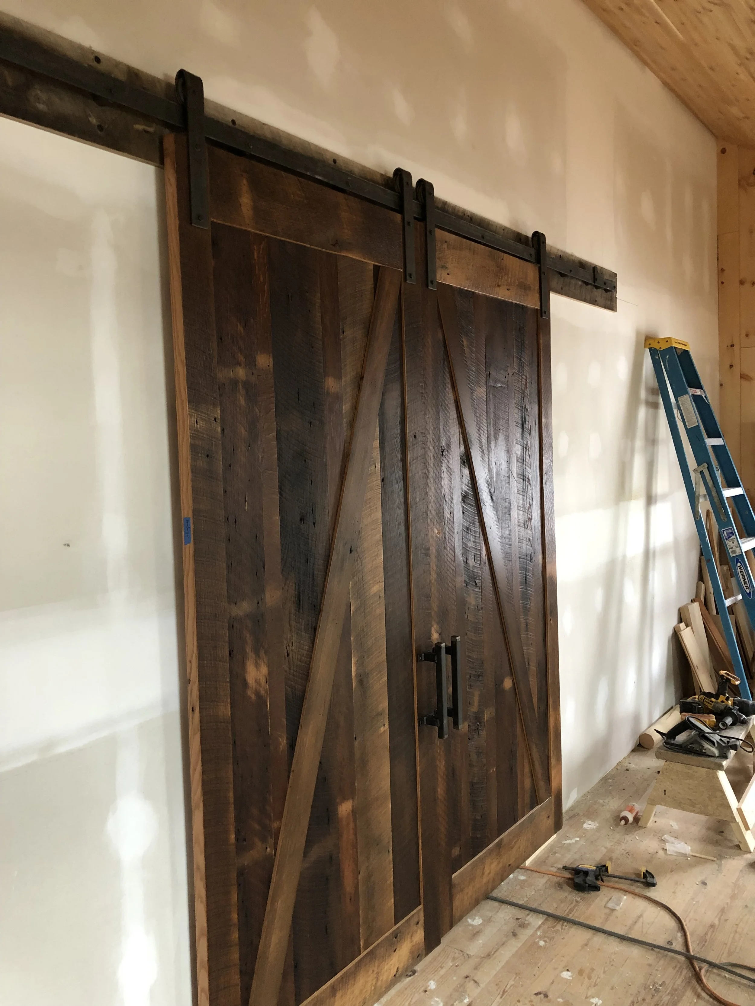 A rustic wooden barn door on a black sliding track, partially open, within a room under construction. Construction tools and a ladder are on the floor and propped against the wall.