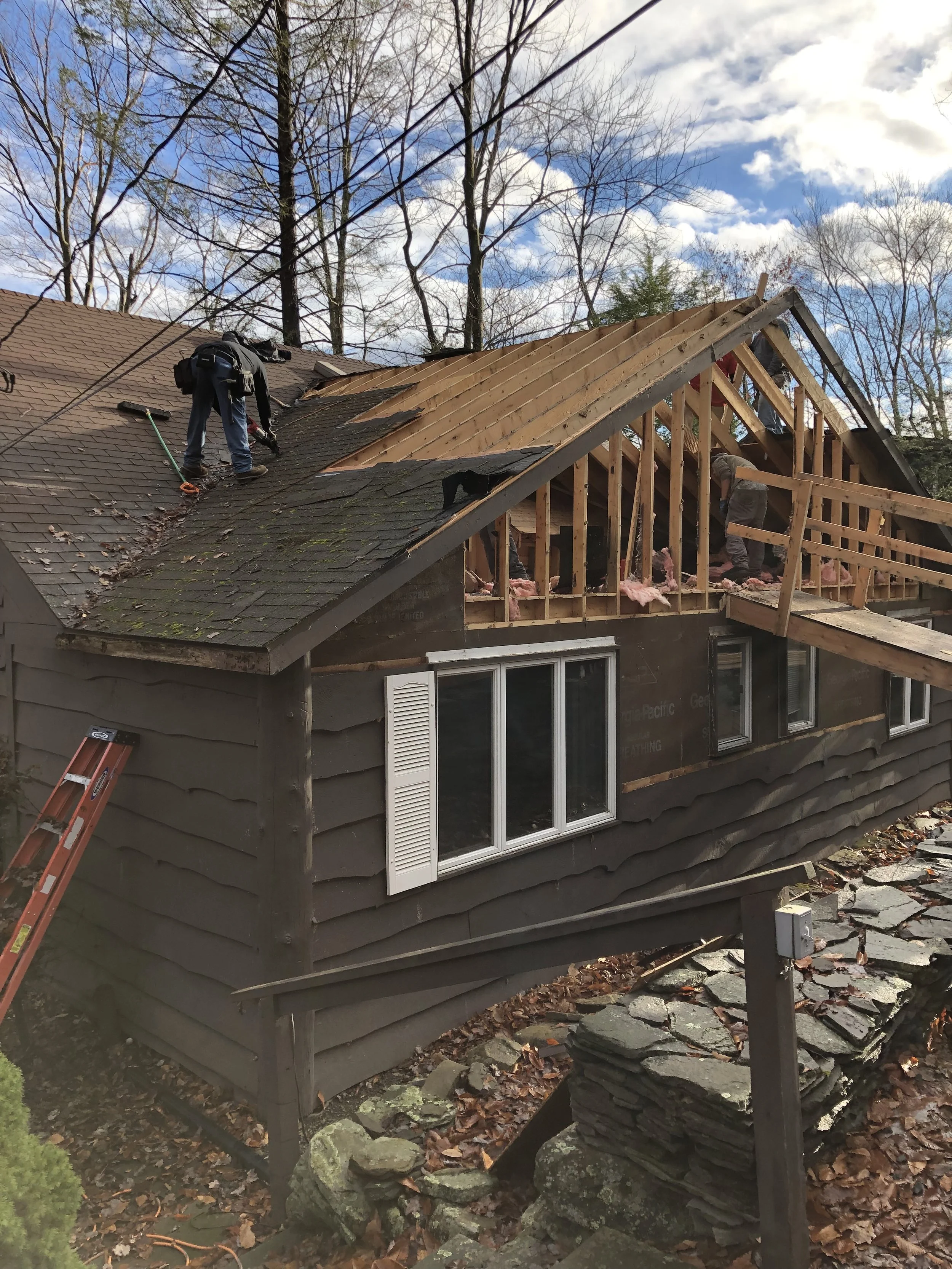 Construction workers replacing or repairing the roof of a house, with part of the roof removed revealing framing and insulation, while tools and equipment are visible on the roof and ladder leaning against the side.