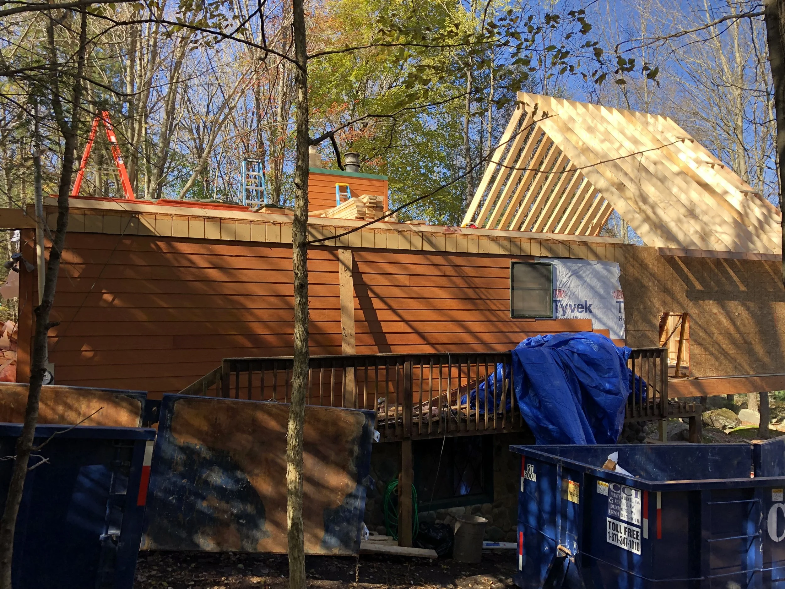 Construction of a house in a wooded area. The house has wood siding, a deck, and a roof with framing in progress. Construction materials and tools are visible around the site.