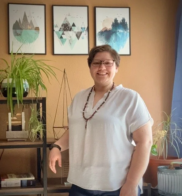 A person with short hair and glasses wearing a white short-sleeve shirt, smiling, standing in a living room with framed art on the wall and plants on a black shelf.