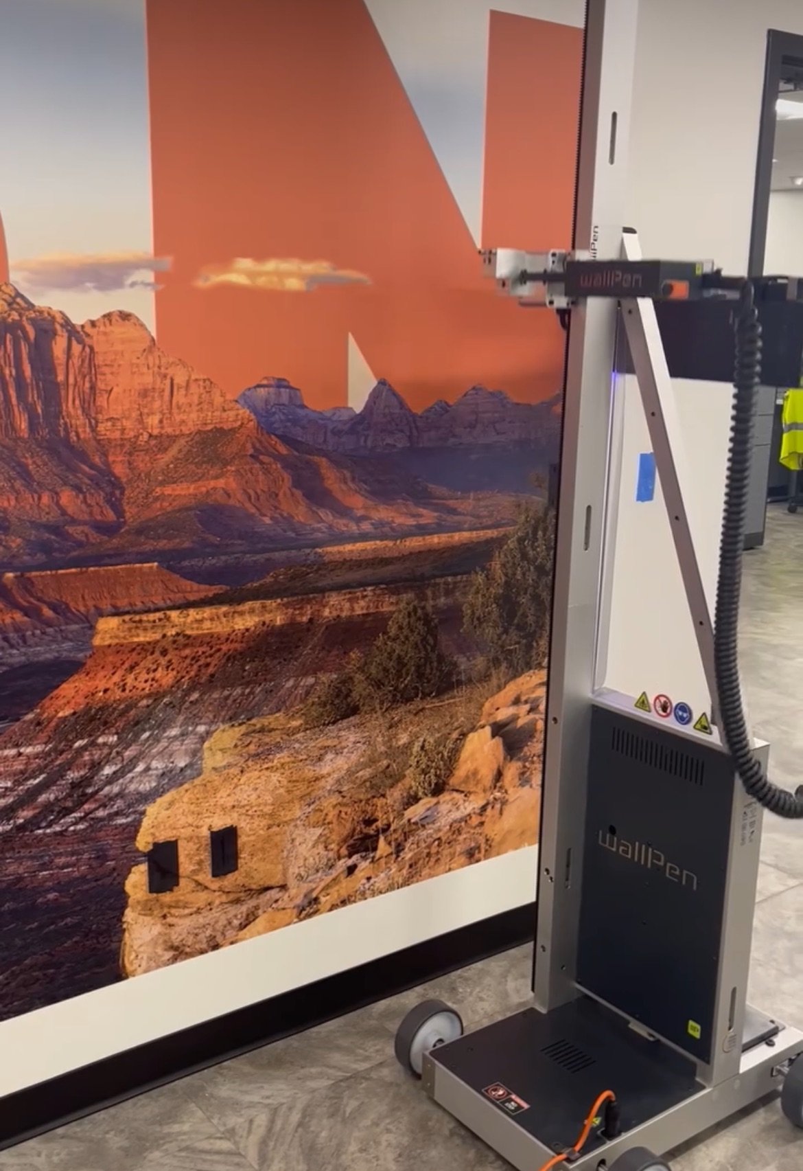 A large printed landscape image of a canyon with mountains in the background, mounted on a stand with a wall printer device called Wallpen positioned in front of it.