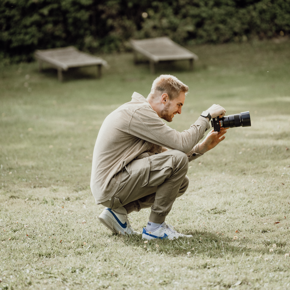 Ein Mann kniet auf der Wiese und fotografiert mit einer Kamera, im Hintergrund sind zwei Liegestühle und Büsche.