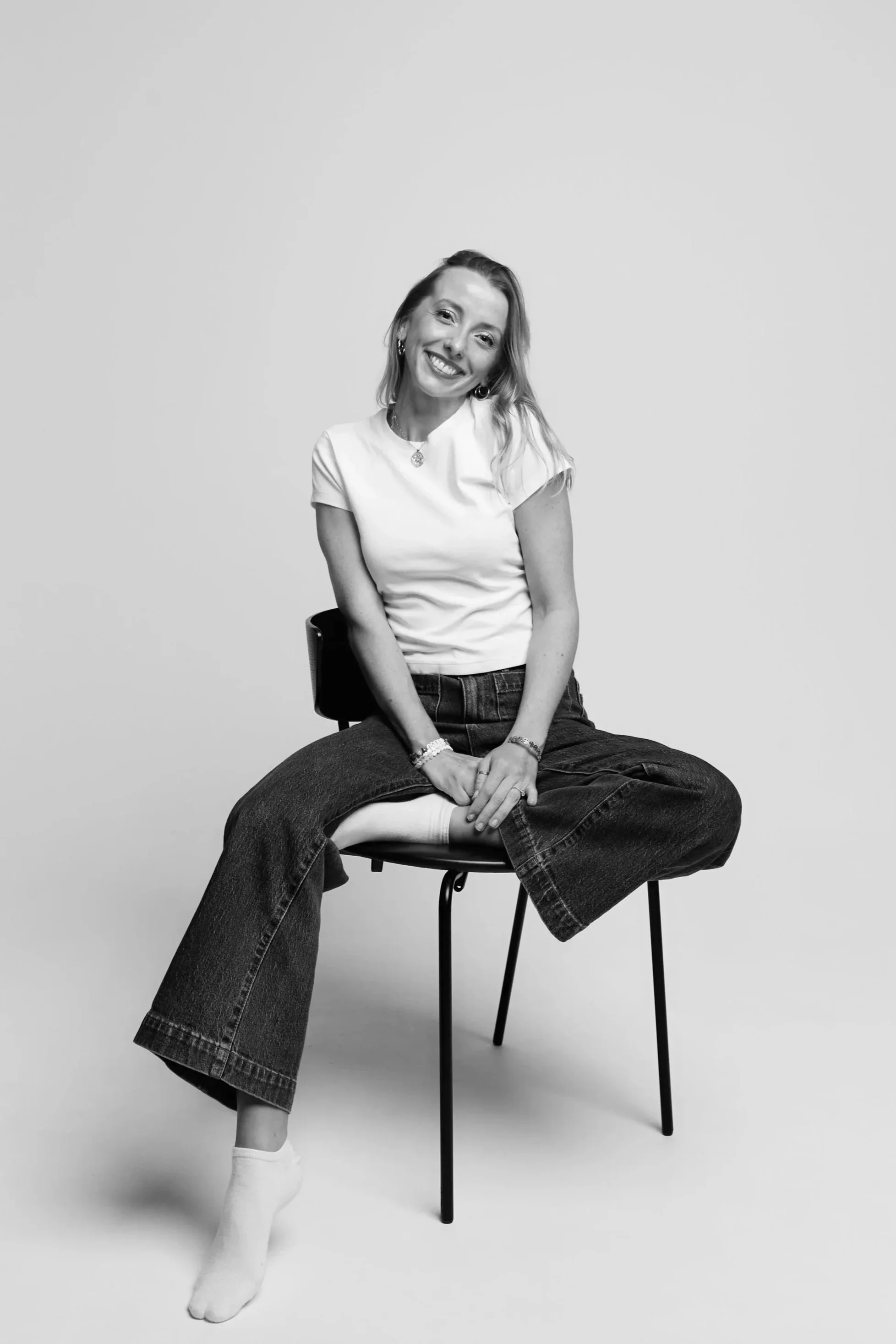 A woman sitting on a chair with her legs crossed, smiling, wearing a white t-shirt, jeans, and socks, in a studio with a plain background.