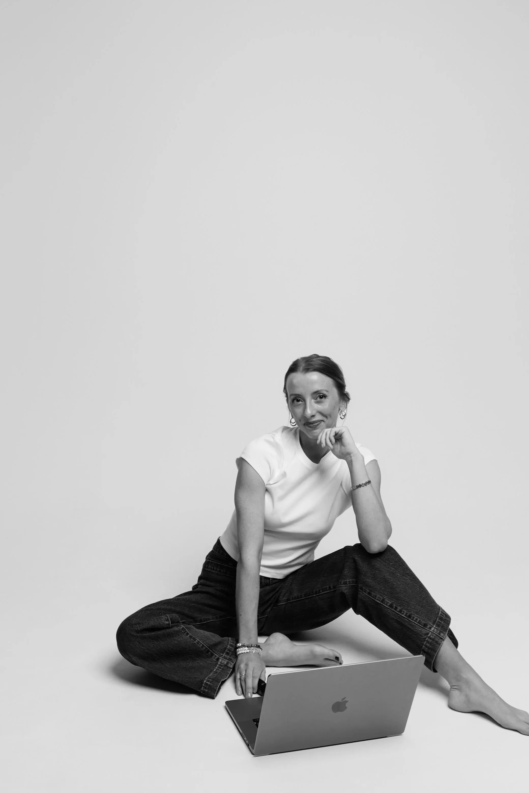 A woman sitting cross-legged on the floor with a laptop in front of her, smiling and resting her chin on her hand, against a plain white background. Coaching clients.