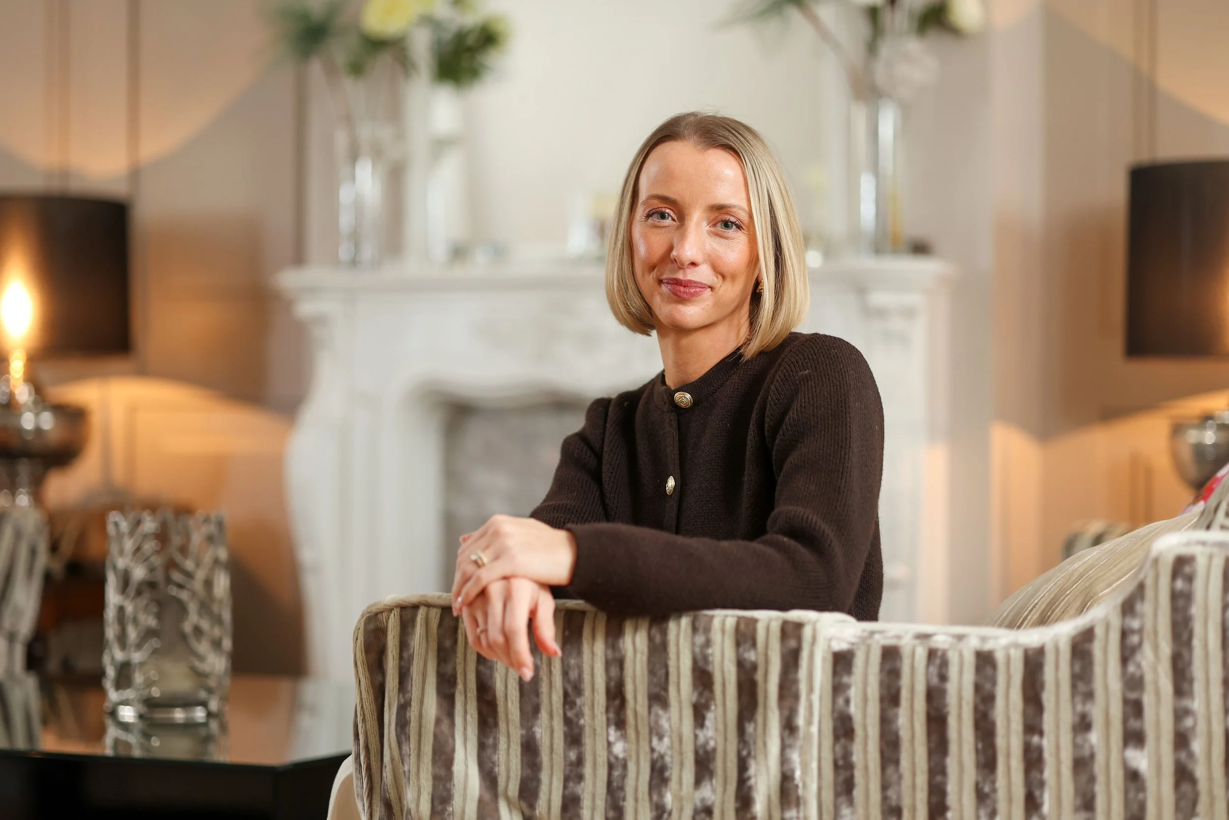 A woman with short blonde hair posing indoors, sitting at a table with a patterned chair in front of her, with blurred table lamps and floral arrangements in the background.