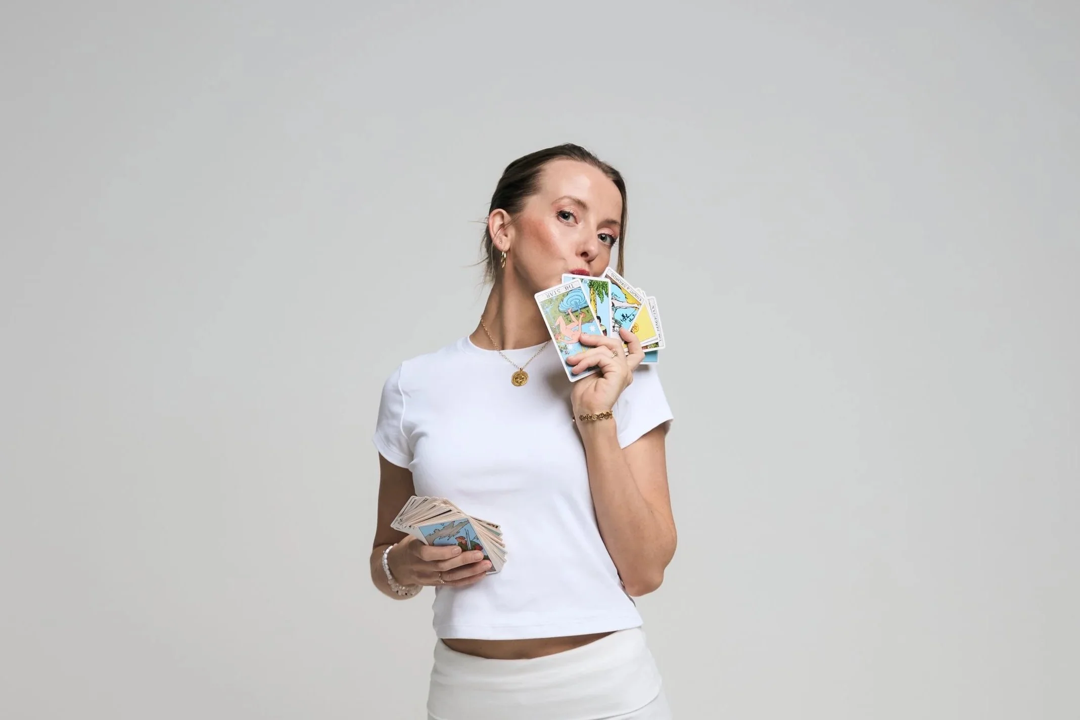 A woman with pale skin and brown hair holding tarot cards near her face, wearing a white t-shirt and jewelry, against a plain light gray background.