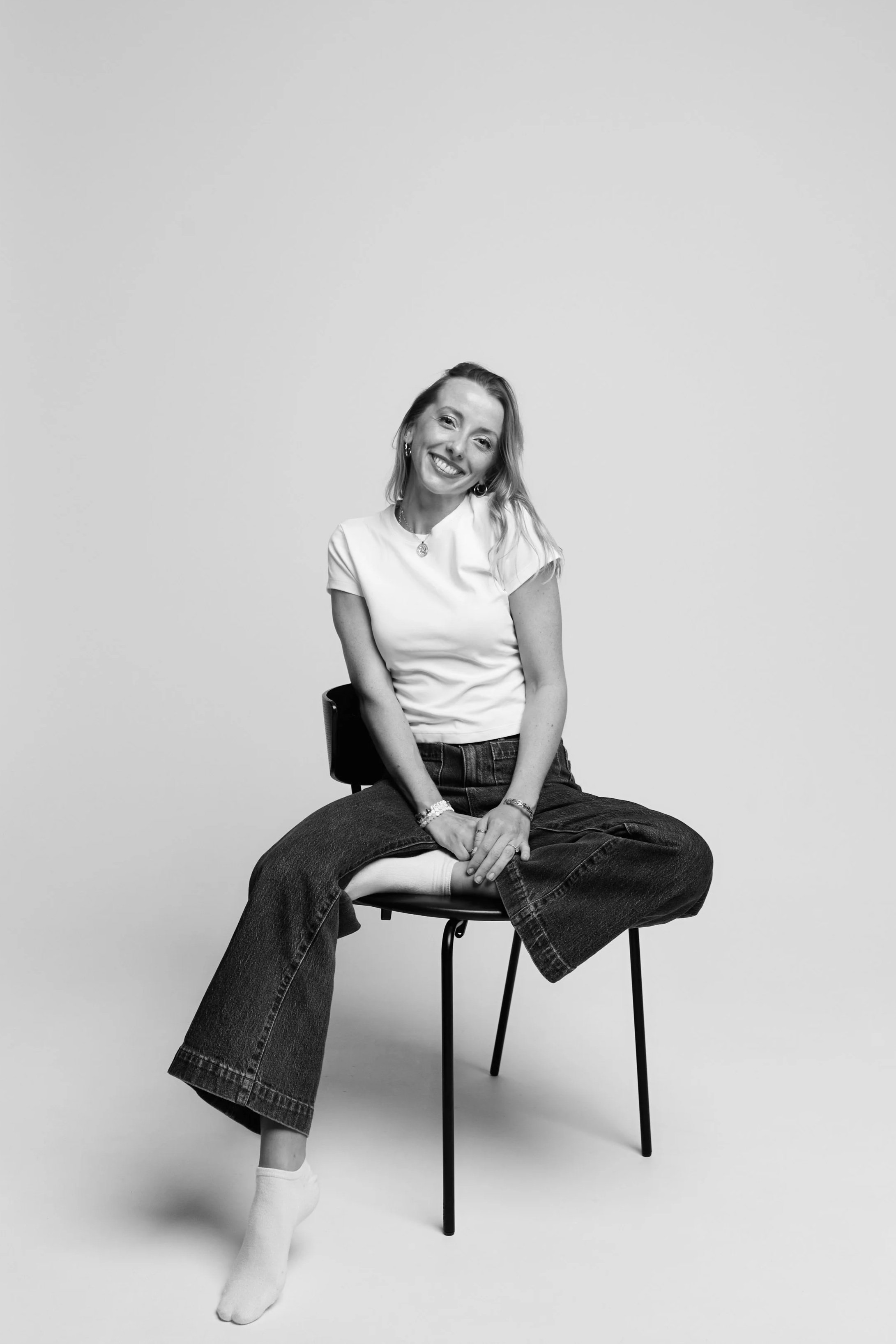 Black and white photo of a woman smiling, sitting on a chair, wearing a short-sleeved shirt, jeans, and socked feet, with her head tilted slightly to the side, against a plain background.