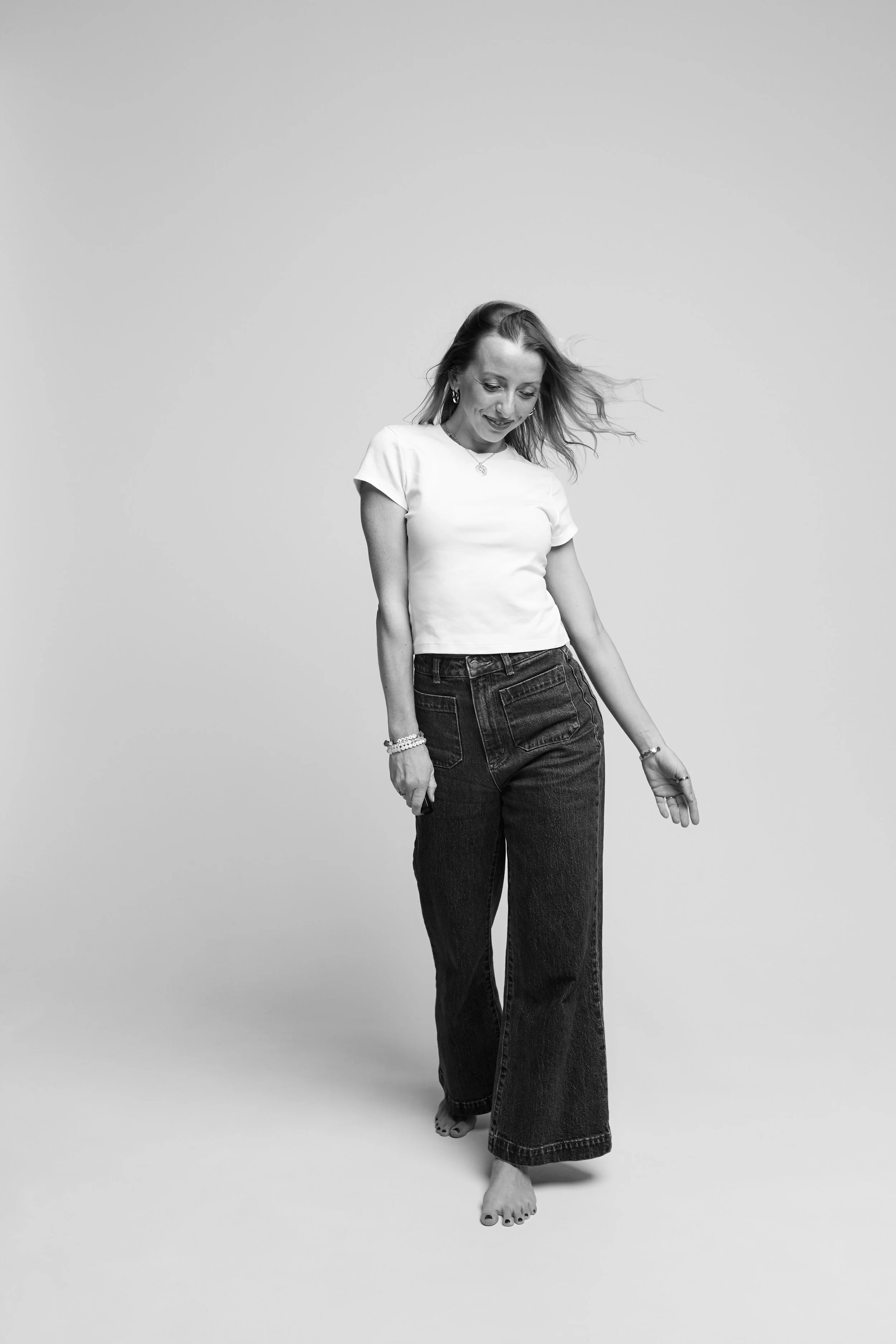 A woman in a white t-shirt and wide-leg jeans standing against a plain light background, looking down and smiling with her hair flowing to the side.