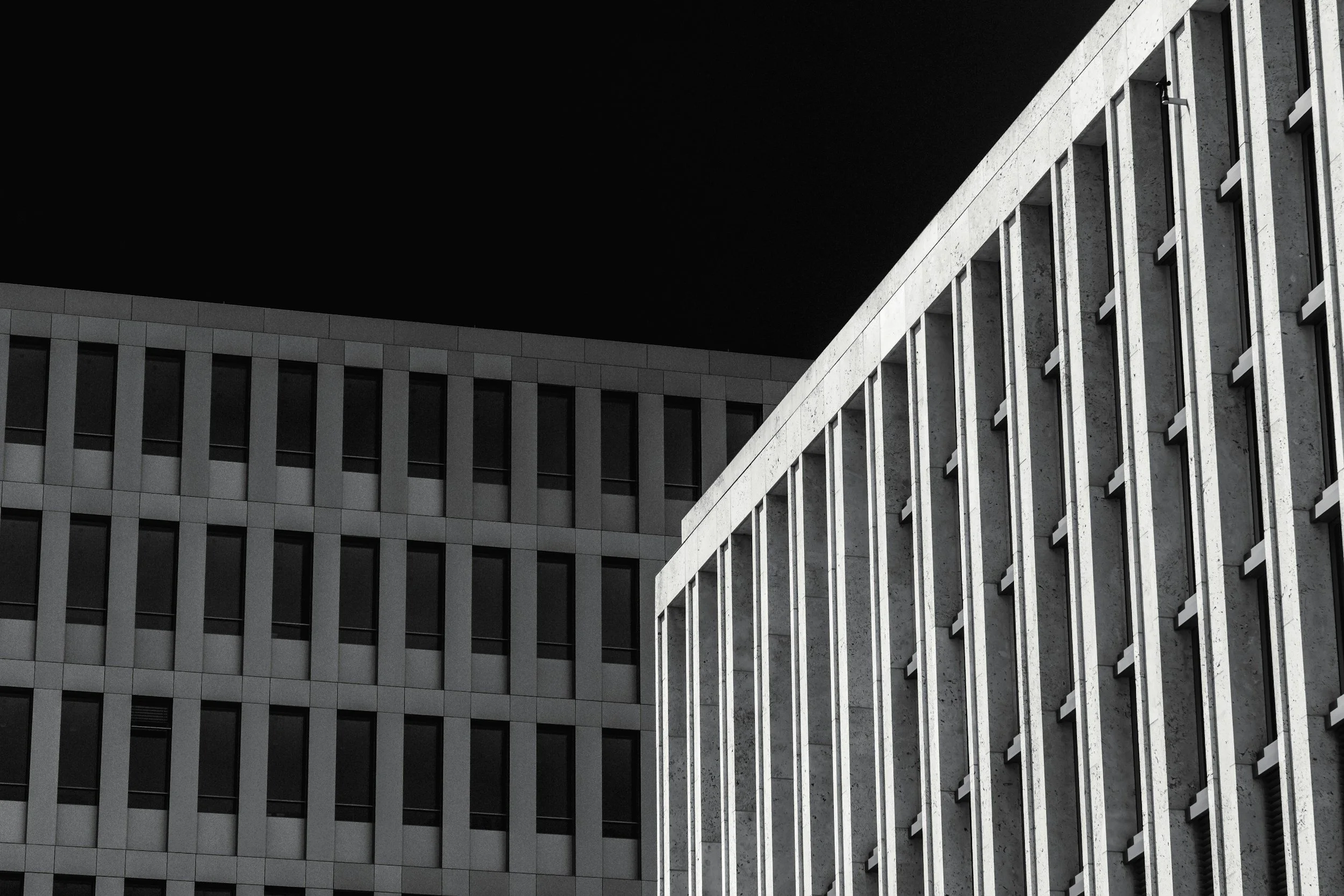 Black and white photo of modern office buildings with grid windows, taken from a low angle at night.