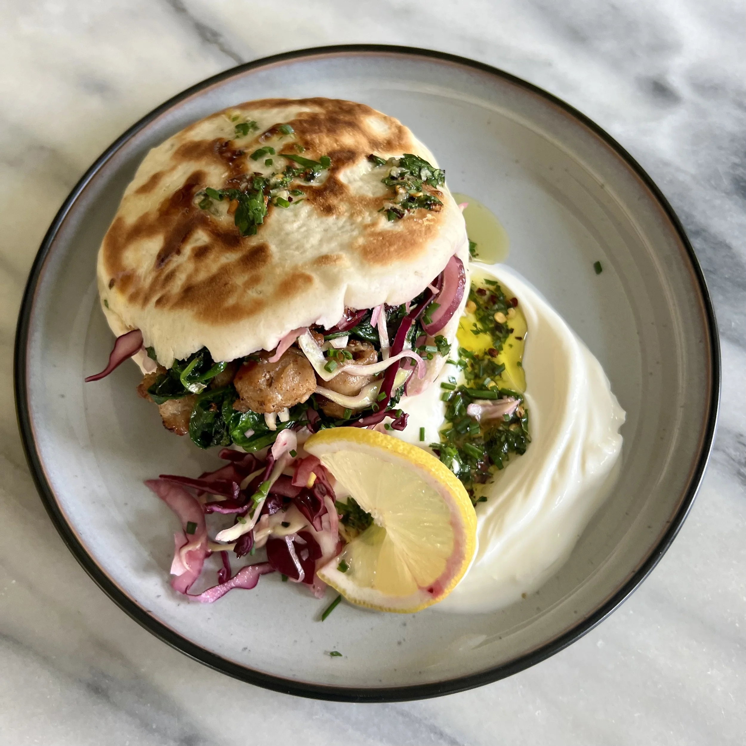 A plate with a sandwich containing greens and meat, topped with chopped herbs and sauce, accompanied by a lemon wedge and a small bowl of sauce or dressing, on a marble surface.