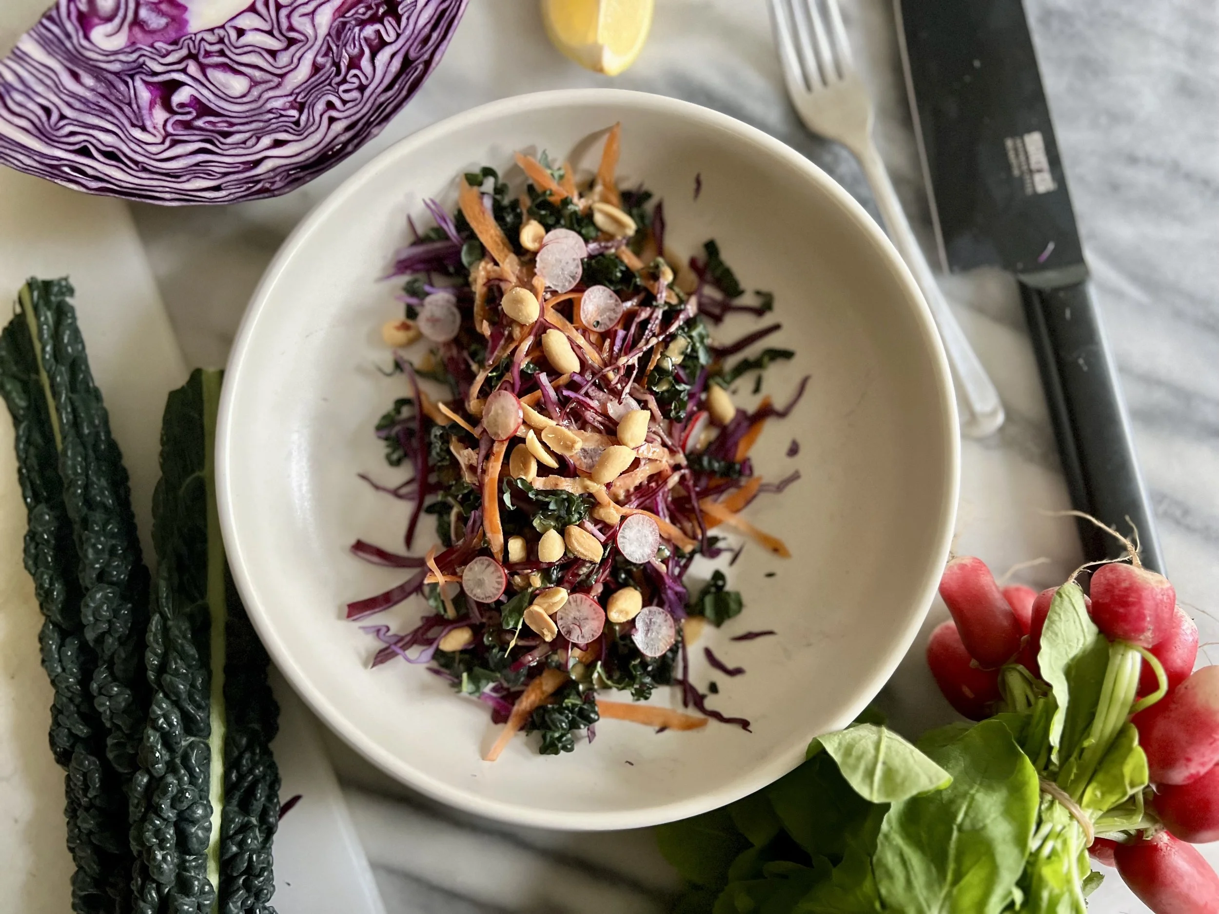 A white bowl of colorful shredded vegetables, including purple cabbage, carrots, radishes, and chopped greens, garnished with pine nuts. Around the bowl are fresh vegetables such as kale, radishes, and a halved purple cabbage, on a light-colored surf