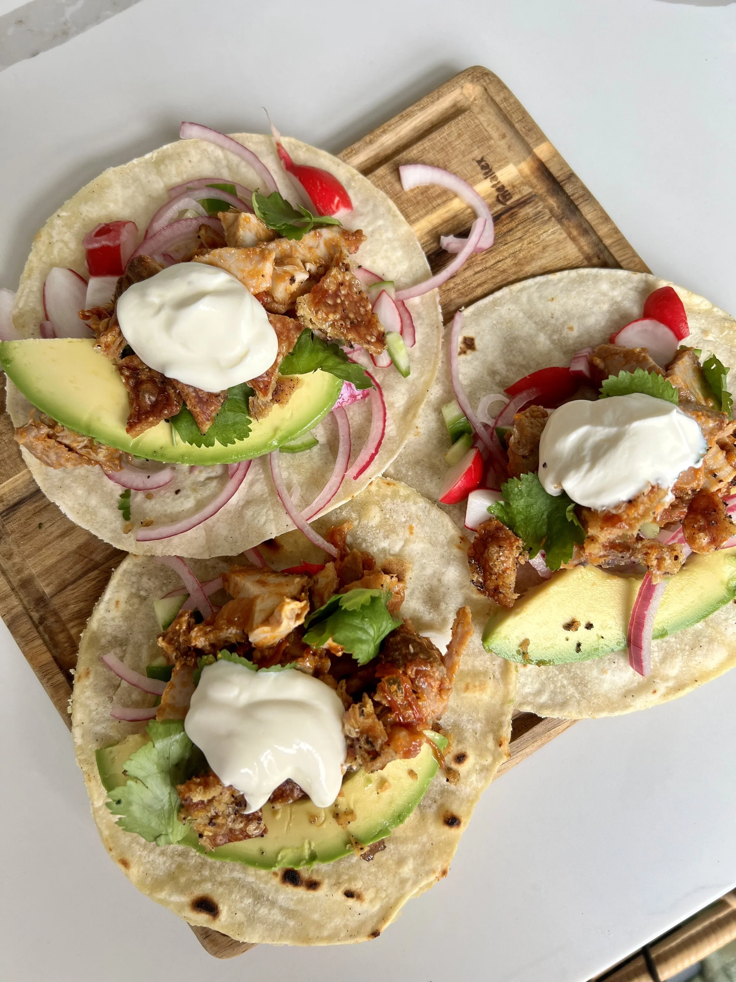Close-up of three tacos on a wooden serving board, filled with shredded meat, avocado slices, radish slices, onions, cilantro, and topped with sour cream.