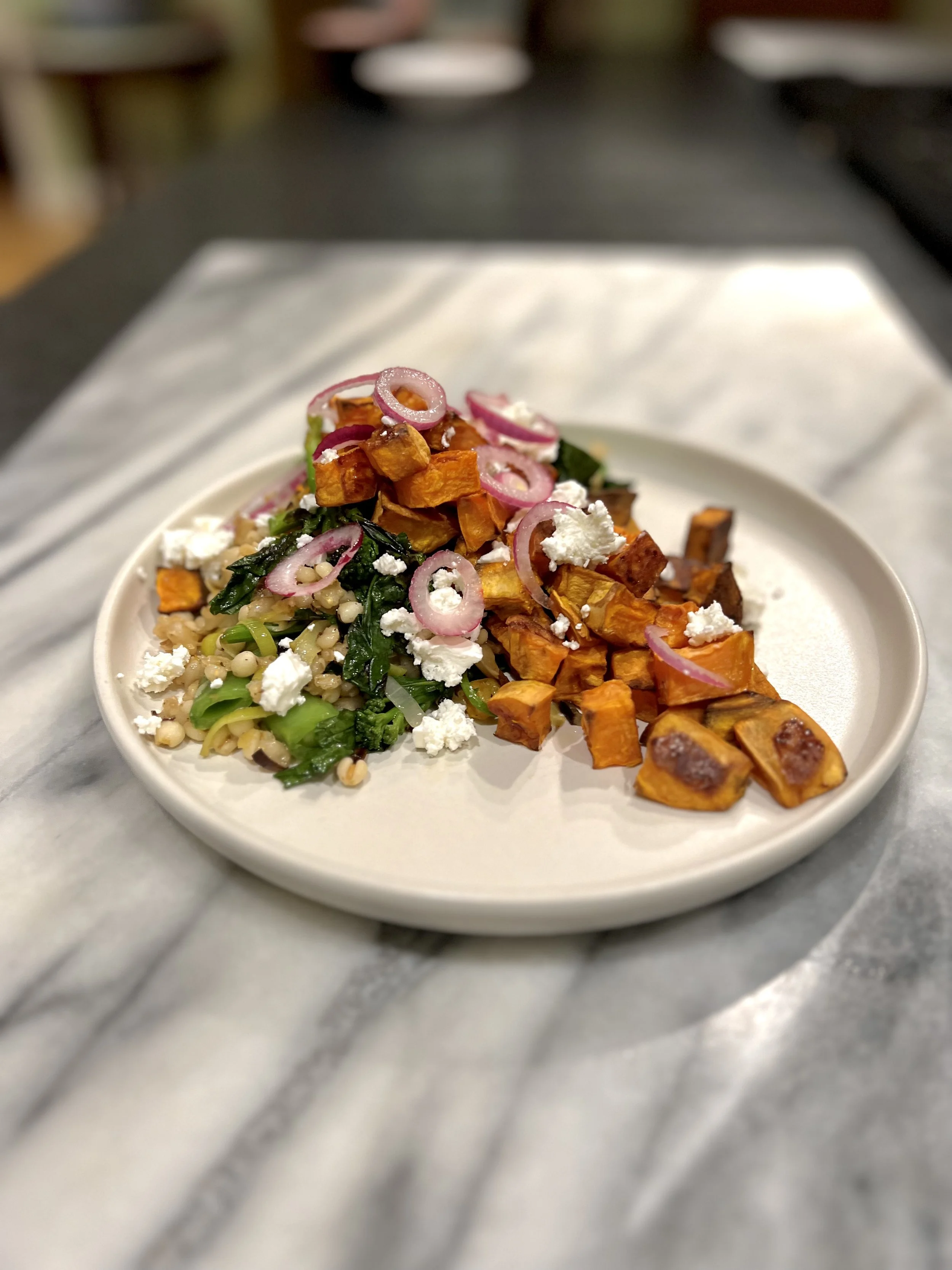 A white bowl containing roasted sweet potatoes, sautéed greens, and a grain salad topped with crumbled cheese and sliced red onions, sitting on a marble countertop.