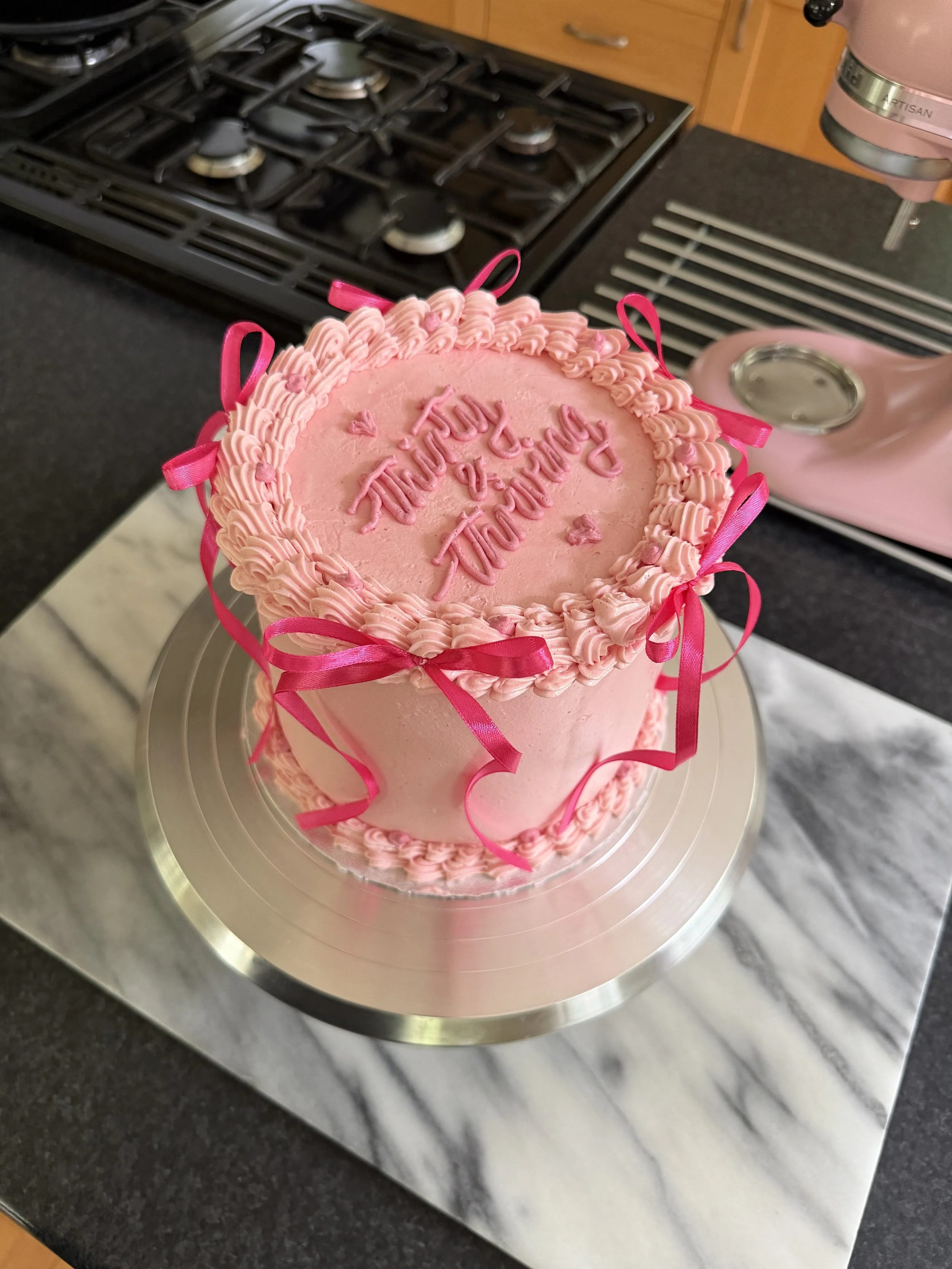 A pink frosted cake with decorative pink ribbons and piped icing, sitting on a silver cake stand in a kitchen.