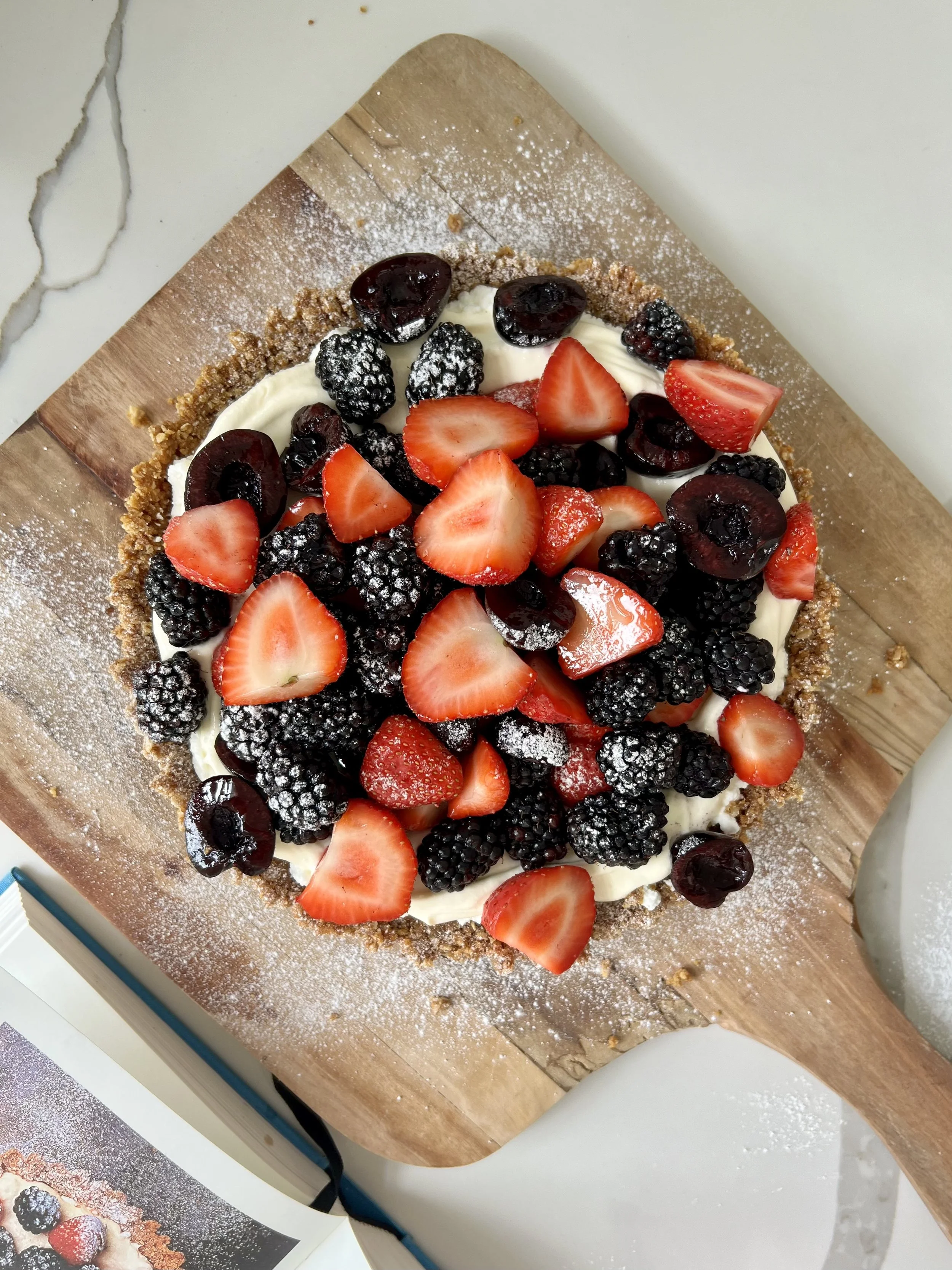 A pie topped with sliced strawberries, blackberries, and cherries, dusted with powdered sugar, on a wooden cutting board.