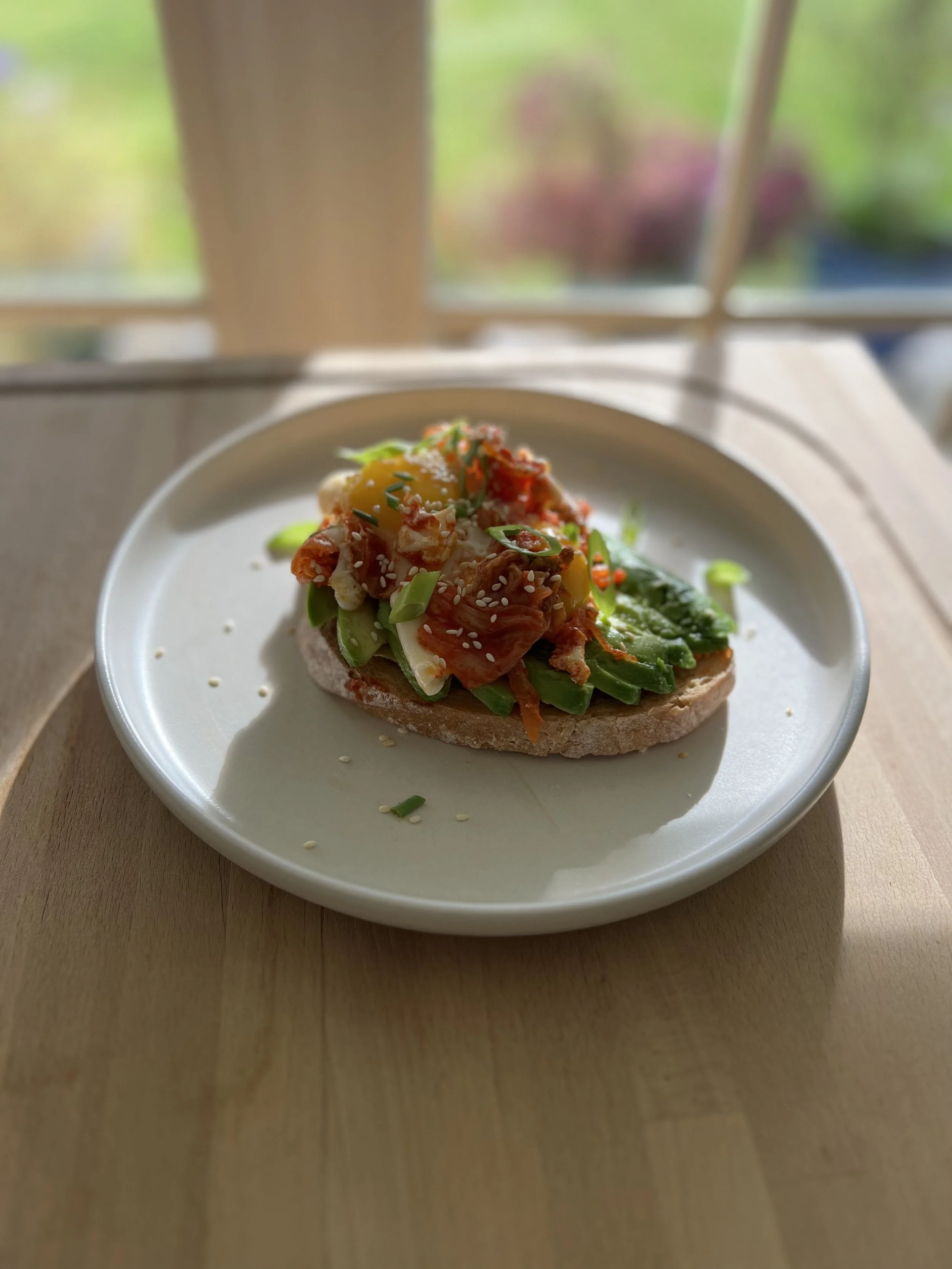 Close-up of an open-faced sandwich on a white plate, topped with vegetables and sesame seeds, with a blurred background of a window and outdoor garden.