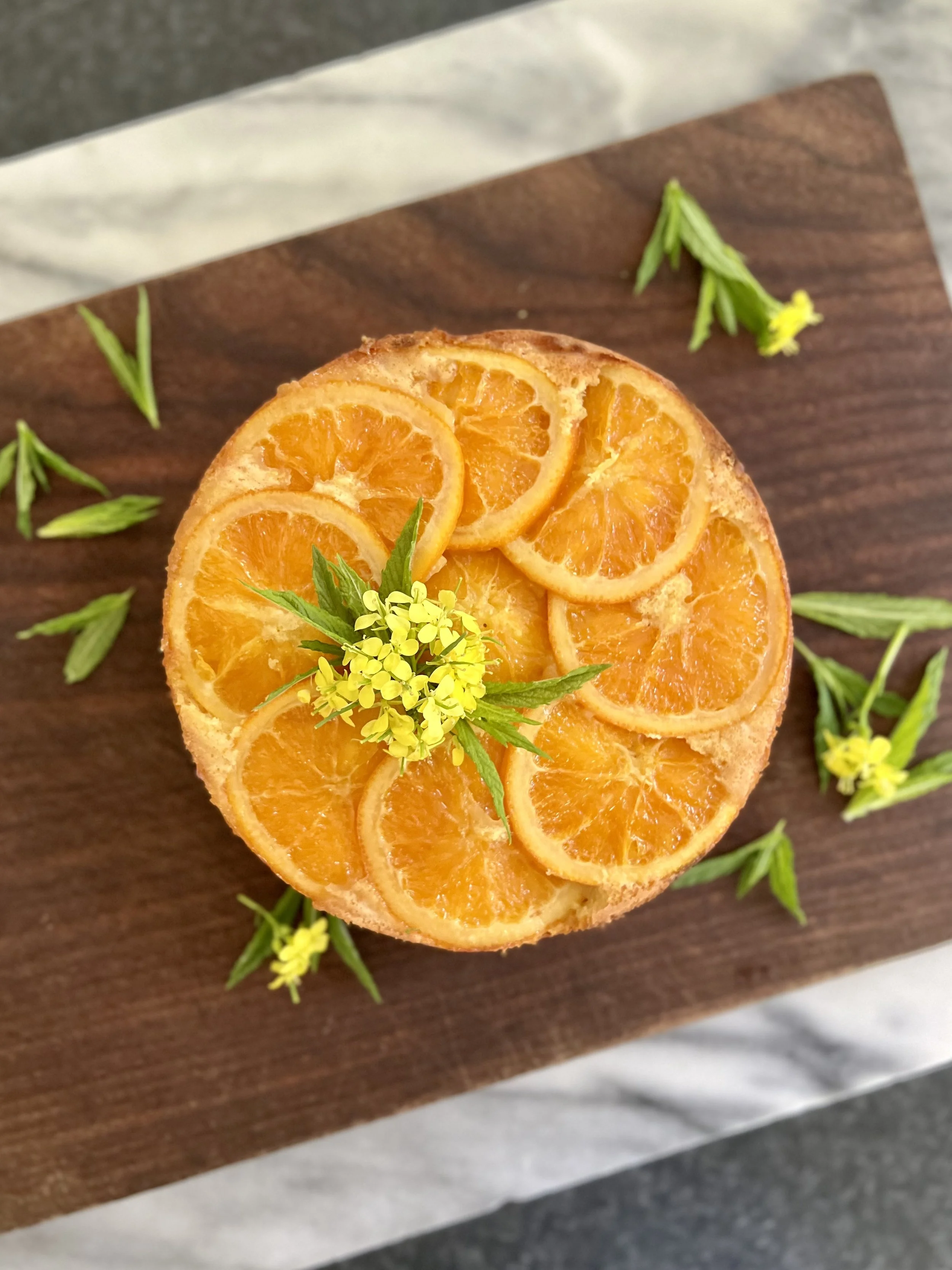 Top view of a round orange cake decorated with sliced oranges and yellow flowers on top, placed on a wooden serving board.