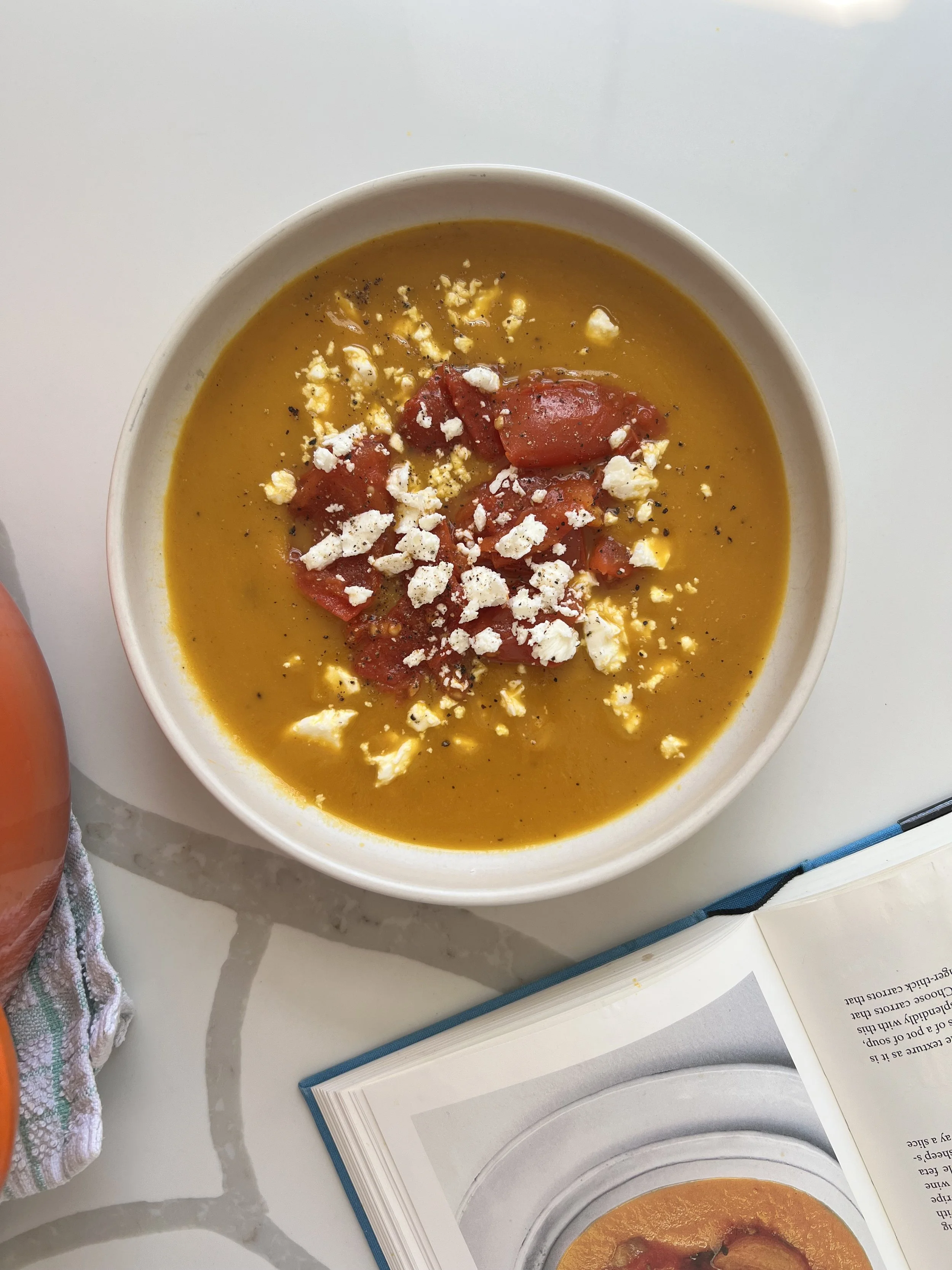 A bowl of pumpkin soup garnished with chopped tomatoes, crumbled white cheese, and black pepper, placed on a white table next to an open cookbook and a striped cloth.