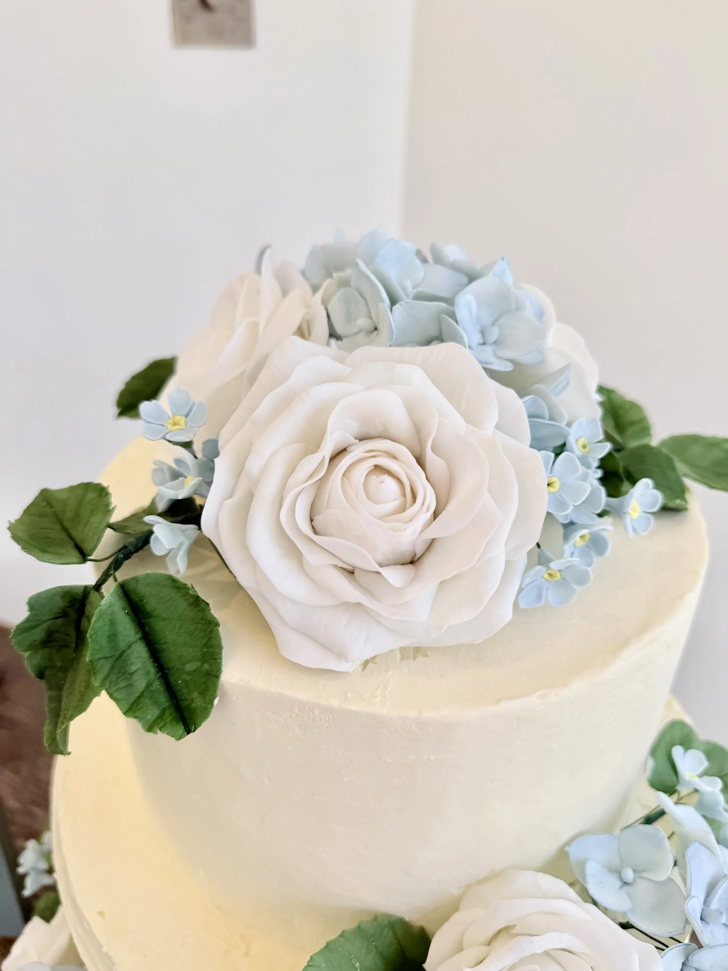 Close-up of a cake topped with realistic-looking white and light blue sugar flowers, including roses and hydrangeas, with green leaves surrounding the flowers.