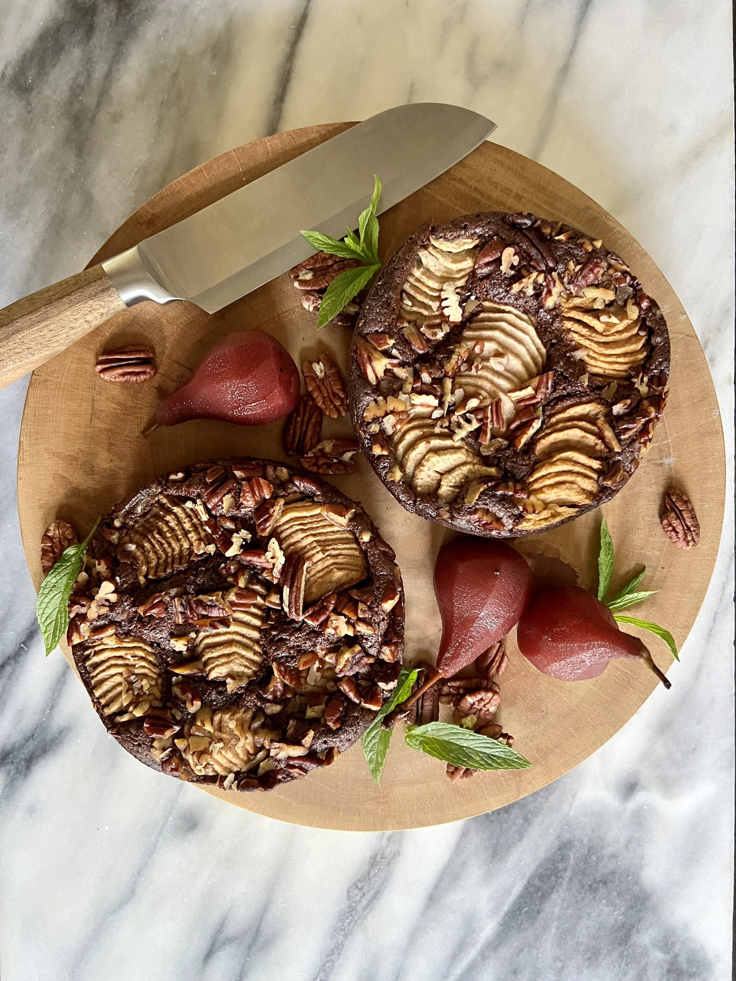 Chocolate pear cake topped with sliced almonds, garnished with fresh mint leaves, on a round wooden serving board with whole pears and additional mint leaves around it, on a marble surface.