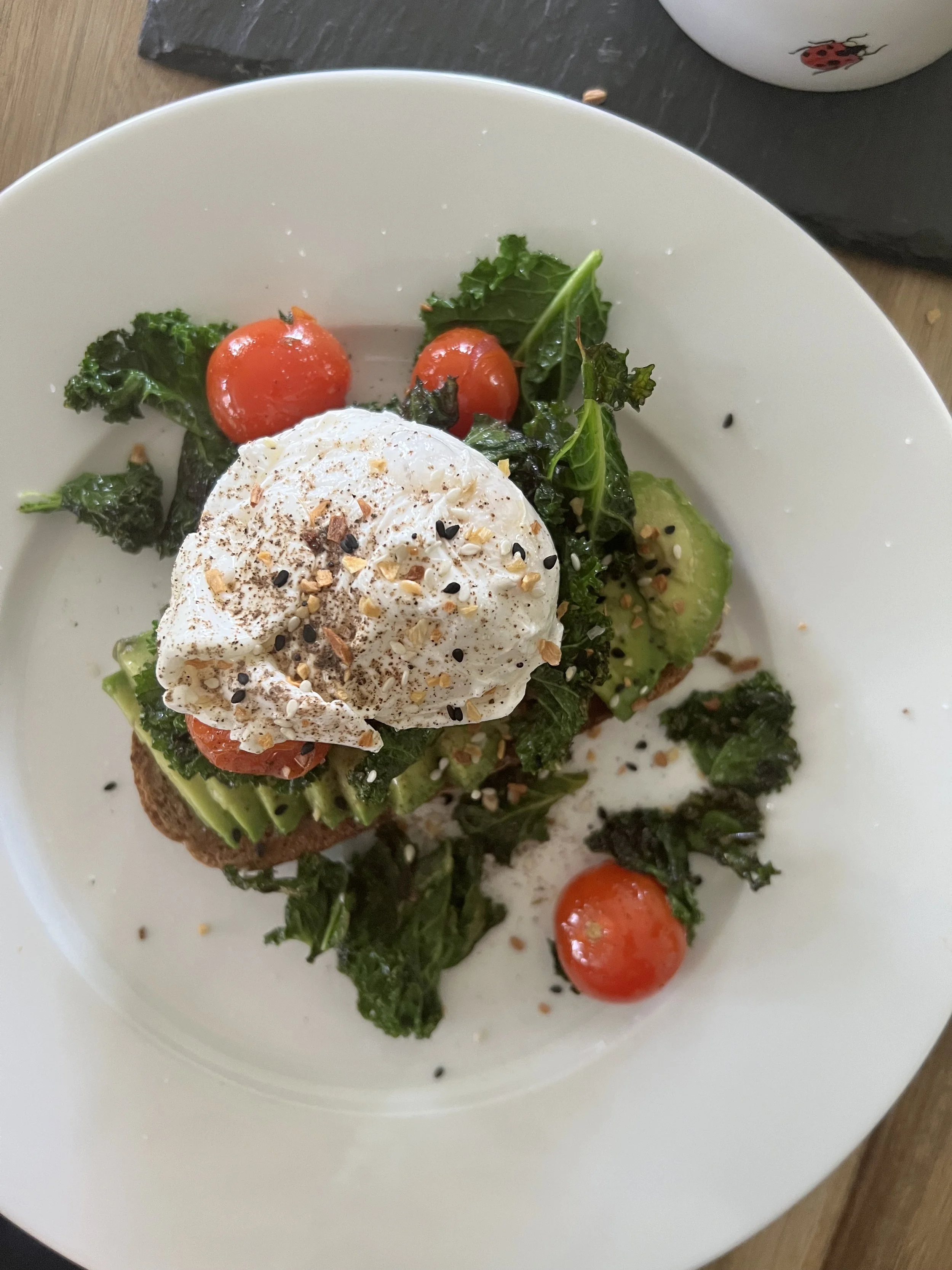 A bowl of salad with cherry tomatoes, kale, avocado slices, and a poached egg sprinkled with black and sesame seeds.