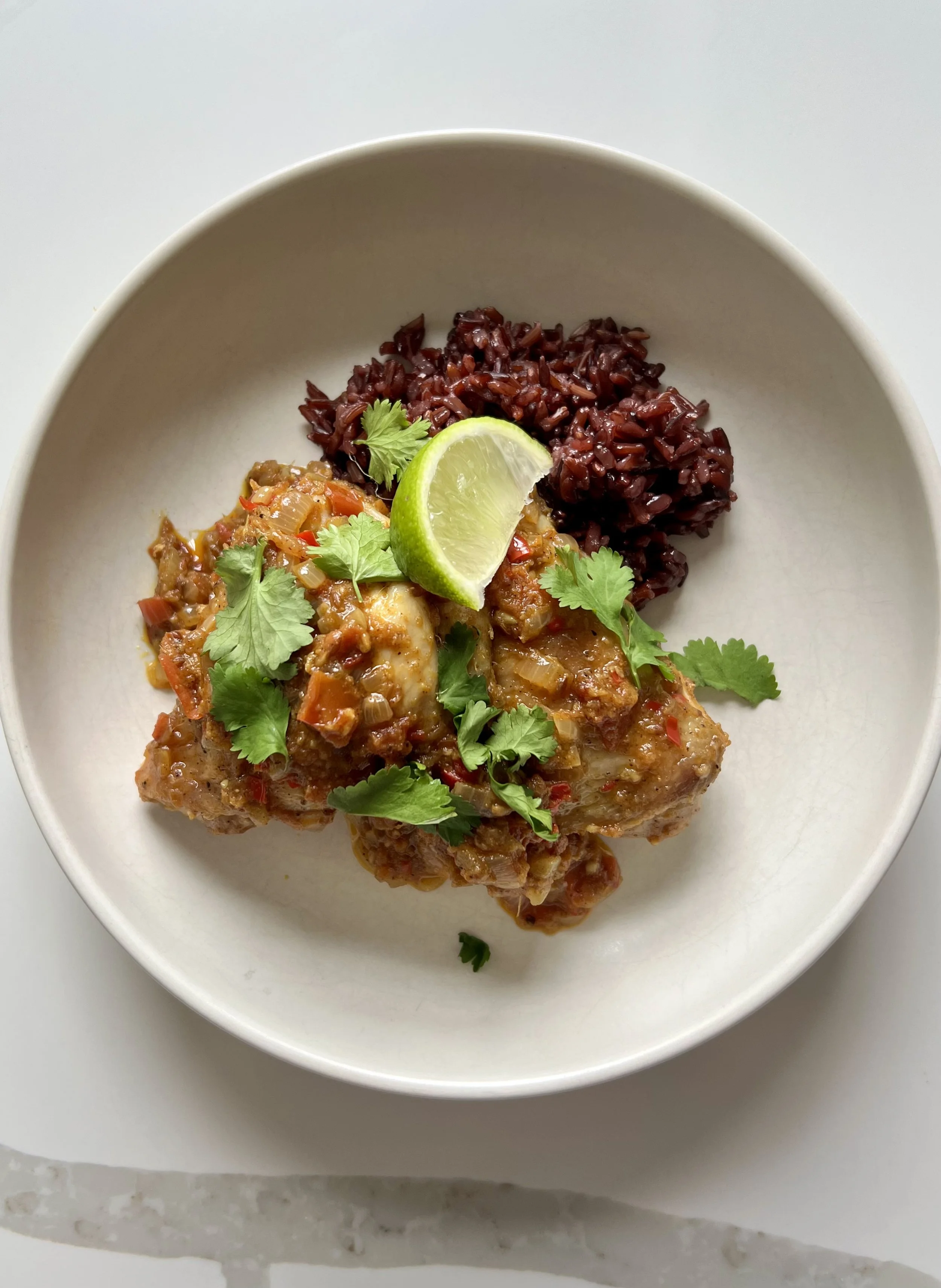 A white bowl with cooked chicken topped with chopped vegetables, garnished with cilantro, a lime wedge, and served alongside cooked black rice.