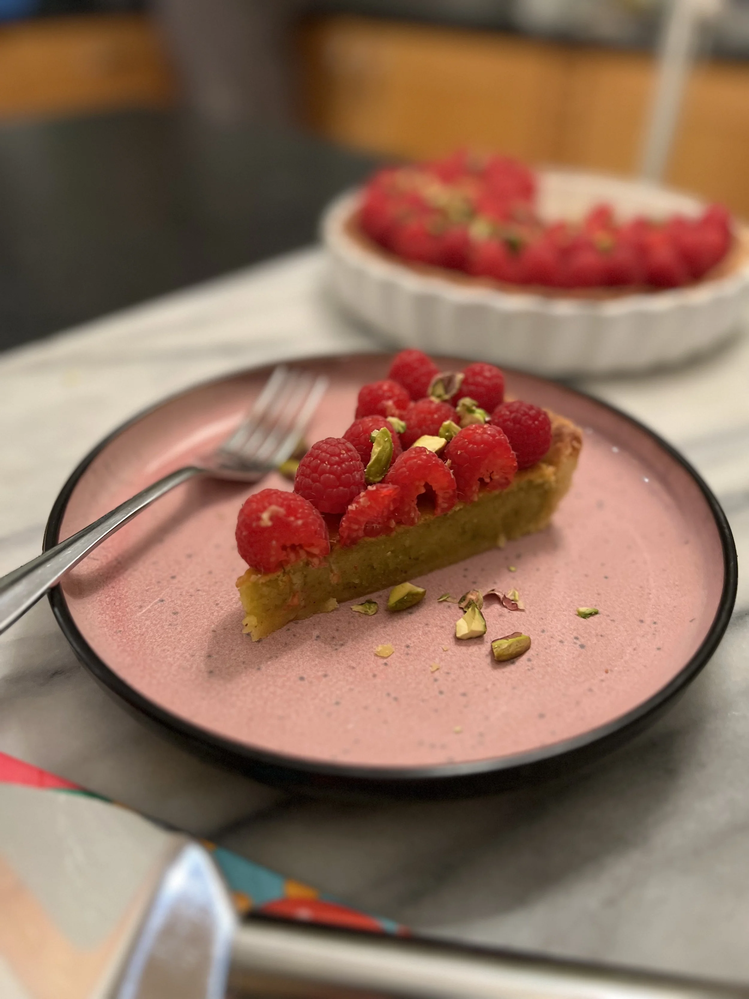 A slice of tart topped with fresh raspberries and chopped pistachios on a pink plate, with a bowl of more raspberries in the background.