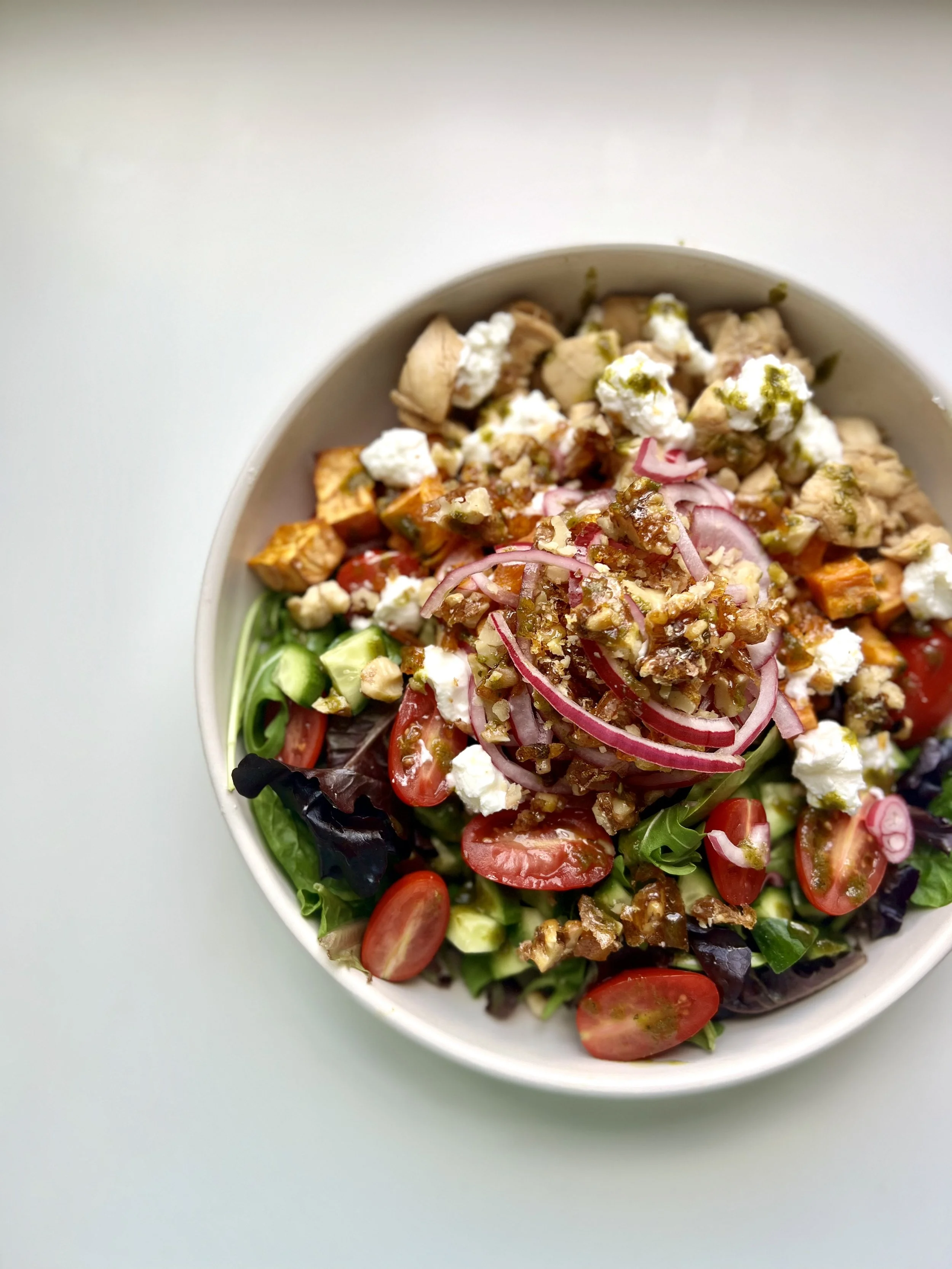 Close-up of a colorful salad in a white bowl, containing cherry tomatoes, cucumbers, red onions, greens, and topped with chicken, feta cheese, and a drizzle of dressing.