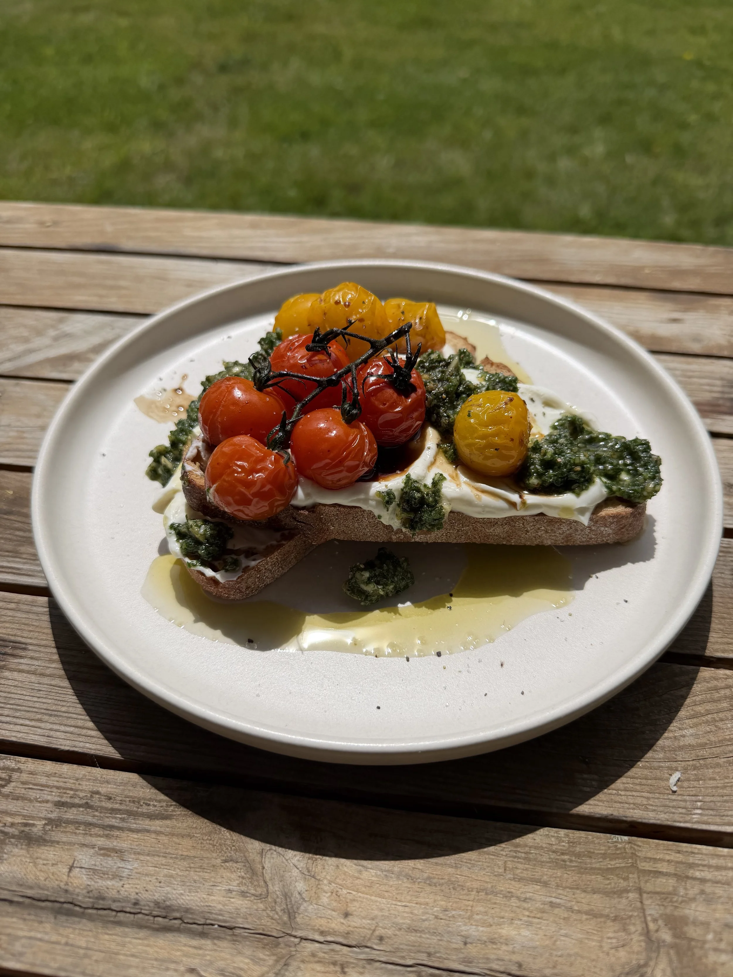Open-faced bread with cherry tomatoes, green pesto, a drizzle of olive oil, and a dollop of cheese on a white plate.