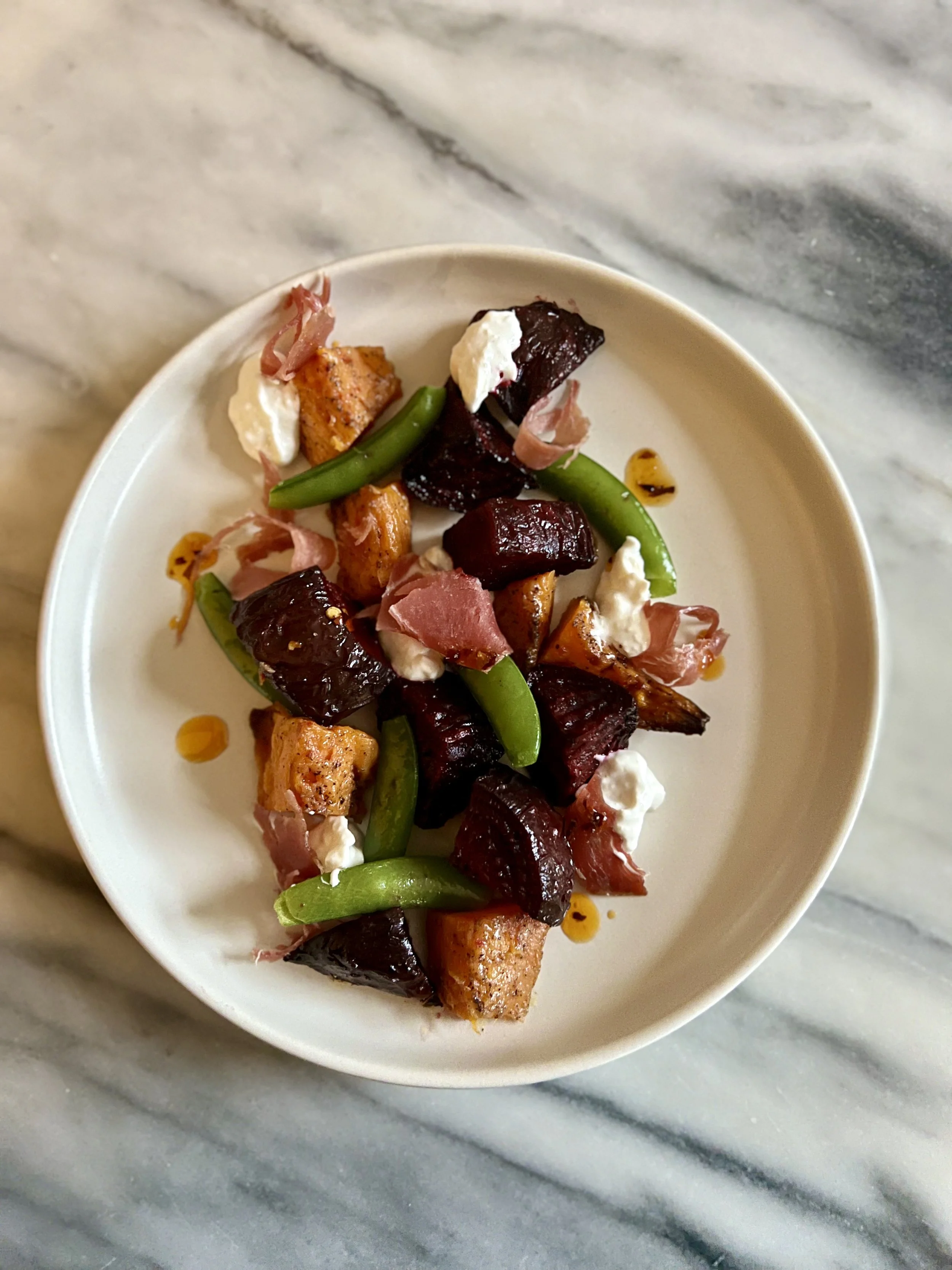 A white bowl of salad containing roasted beets, green snap peas, chunks of grilled chicken, dollops of white cheese or cream, and small pieces of prosciutto, with a drizzle of olive oil, on a marble surface.
