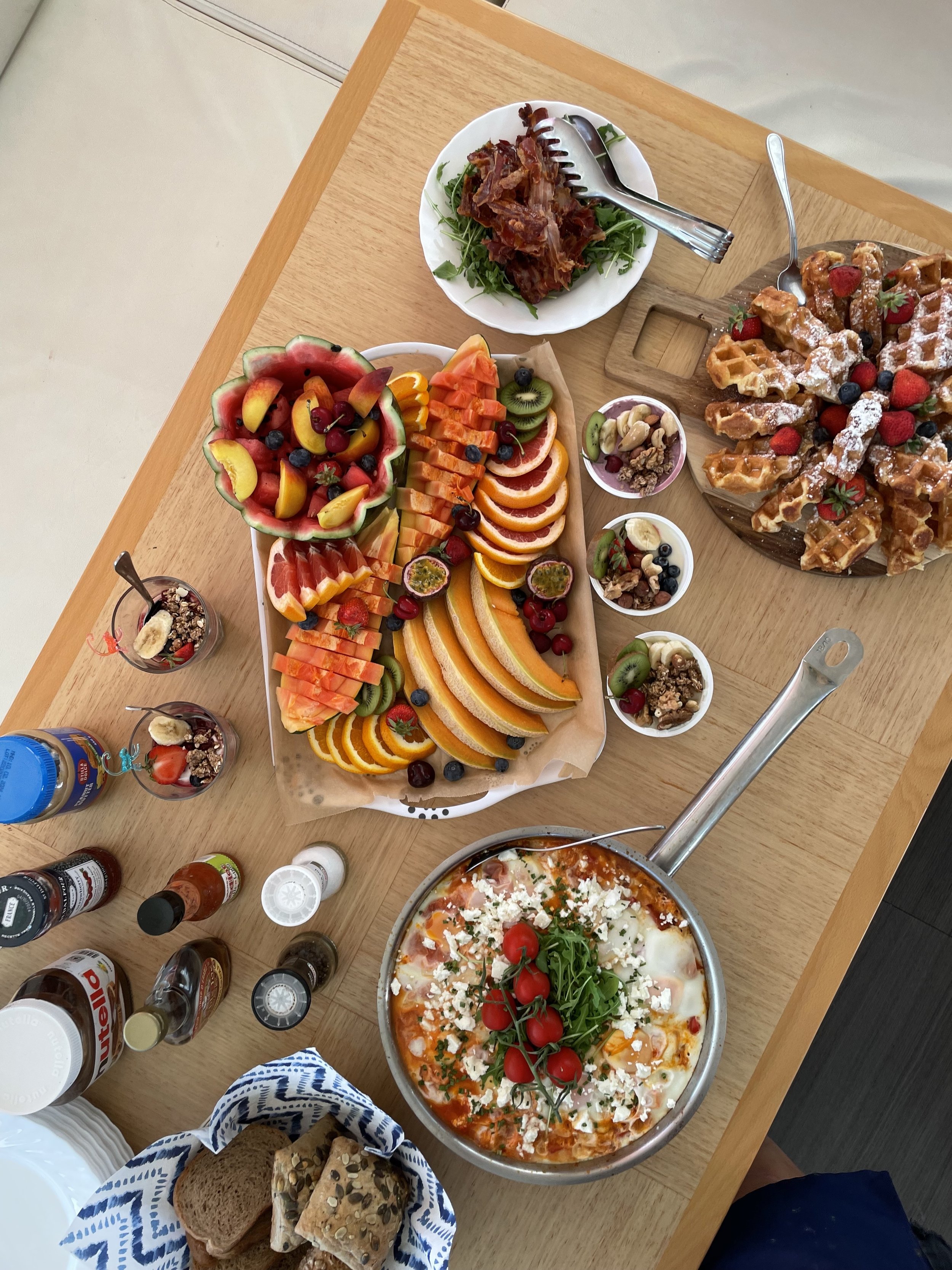 A wooden table with a variety of food including a large fruit platter with watermelon, honeydew, cantaloupe, oranges, grapes, strawberries, and kiwi; a bowl of waffles topped with strawberries and powdered sugar; a pizza with cherry tomatoes, green o