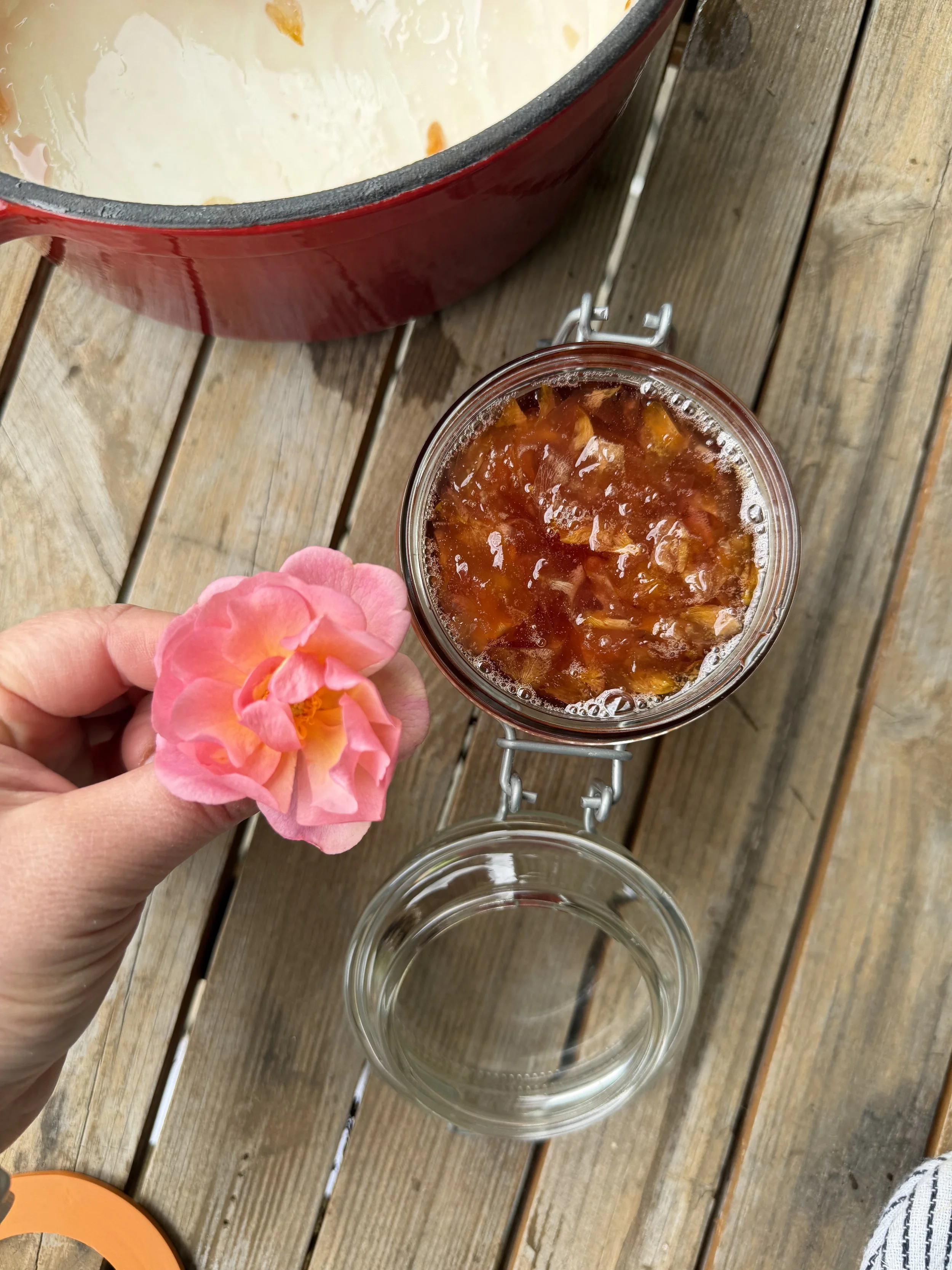 A hand holding a pink flower above a glass jar filled with orange-colored jam or preserve, on a wooden surface. There is a red pot partially visible at the top of the image.