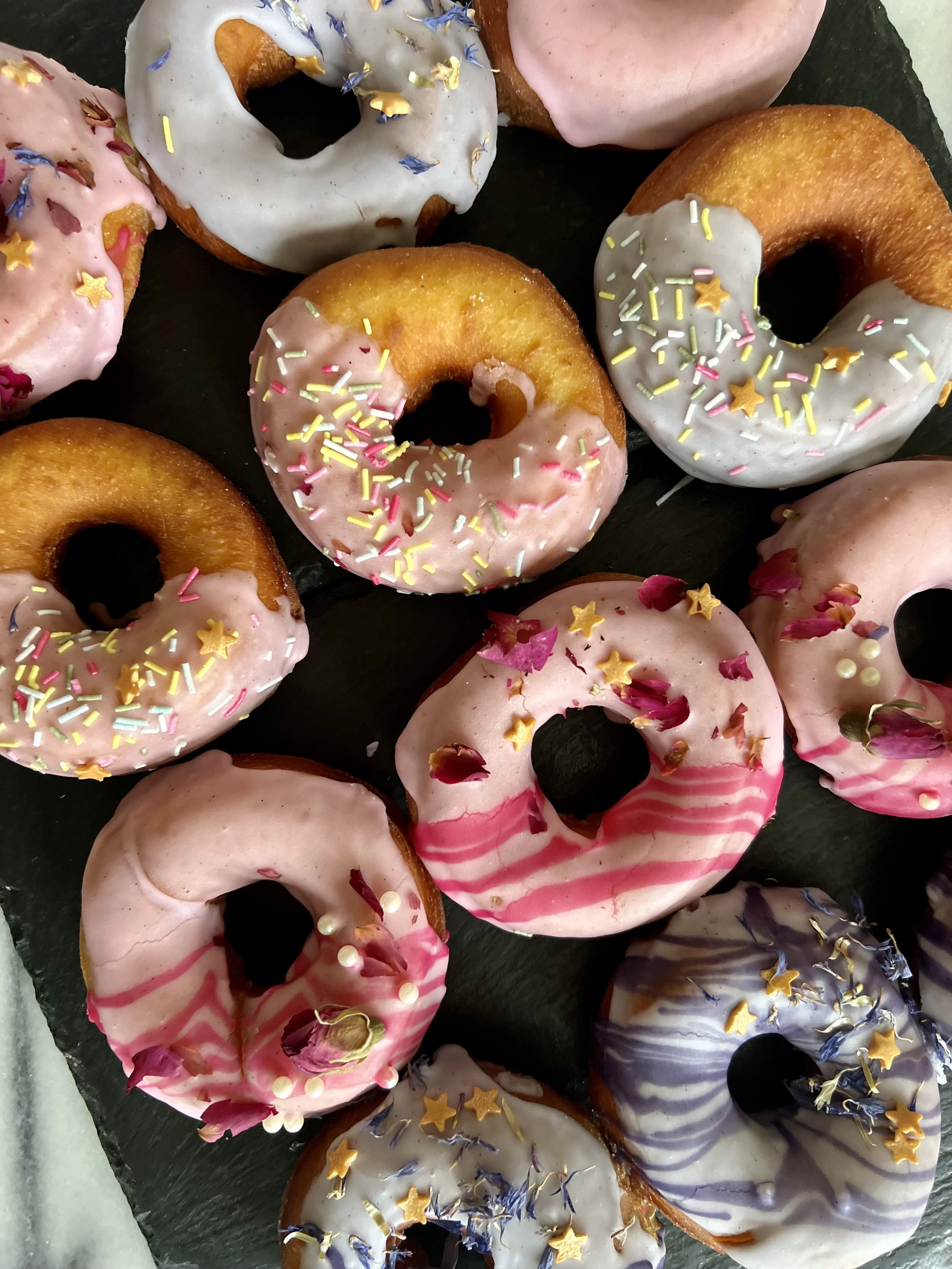 Assorted donuts with pink, white, and purple icing decorated with colorful sprinkles, edible glitter, and dried flower petals, arranged on a black slate serving tray.