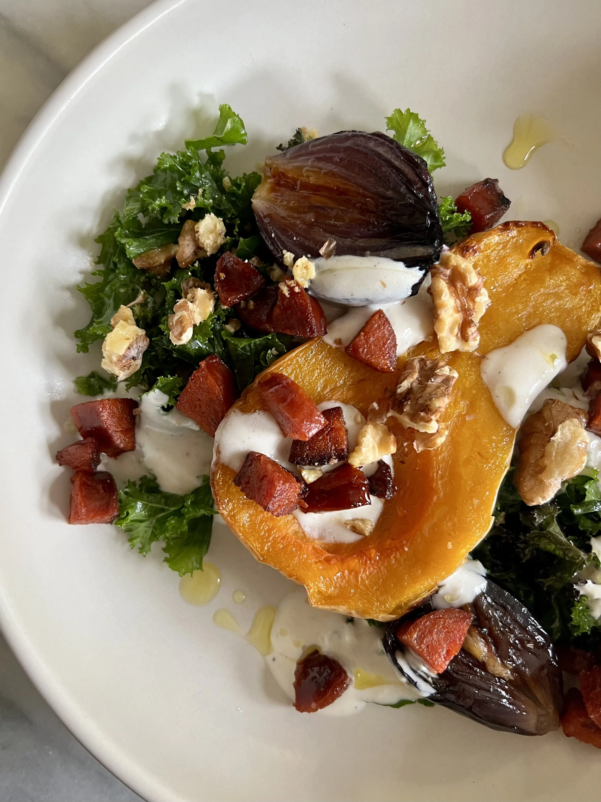 A plate featuring roasted eggplant topped with a creamy sauce, surrounded by chopped bacon, roasted red peppers, chopped walnuts, chopped greens, and drizzled with olive oil.