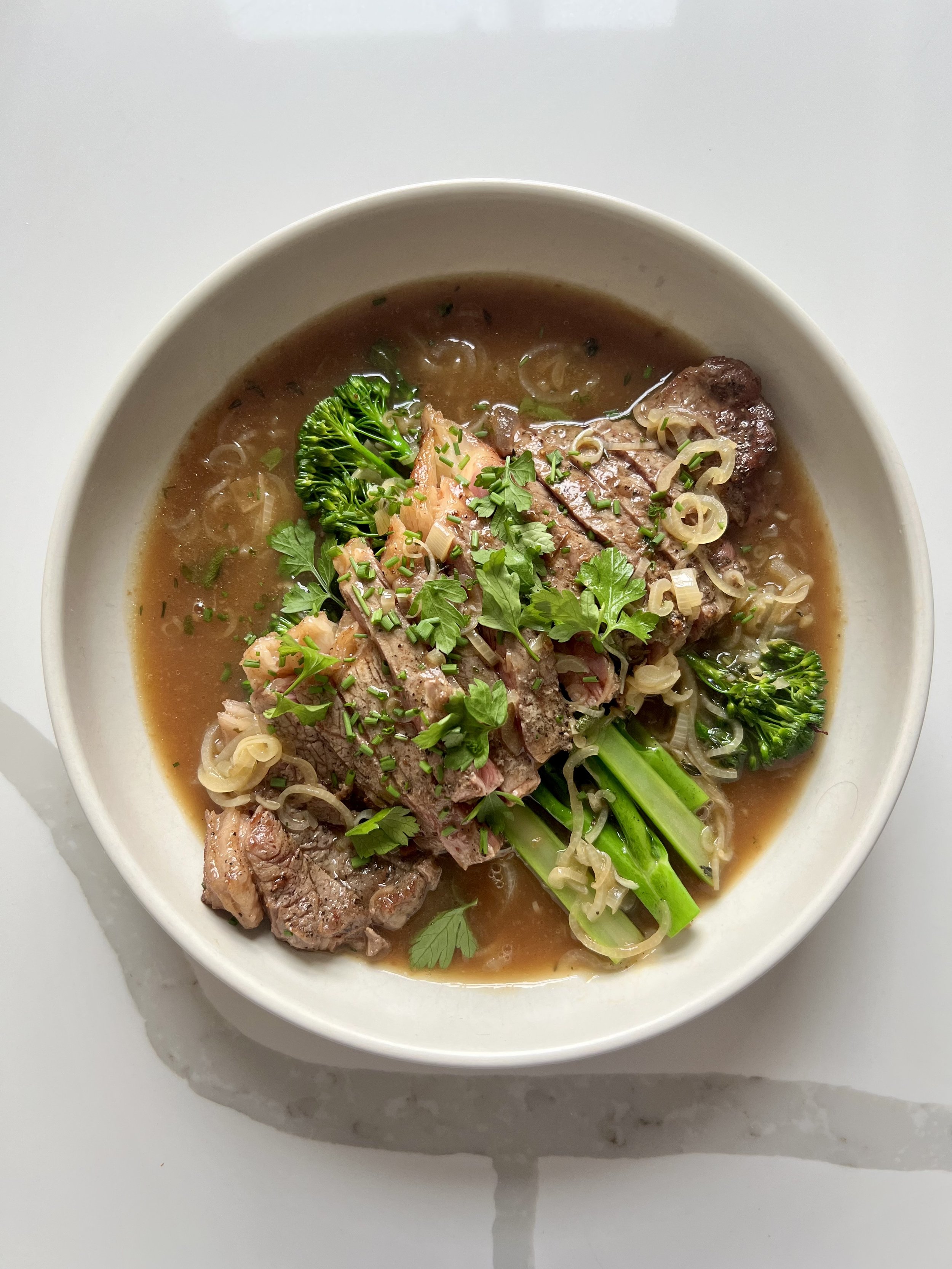 A bowl of beef soup with vegetables, including broccoli, chopped green onions, and fresh herbs, in a broth.